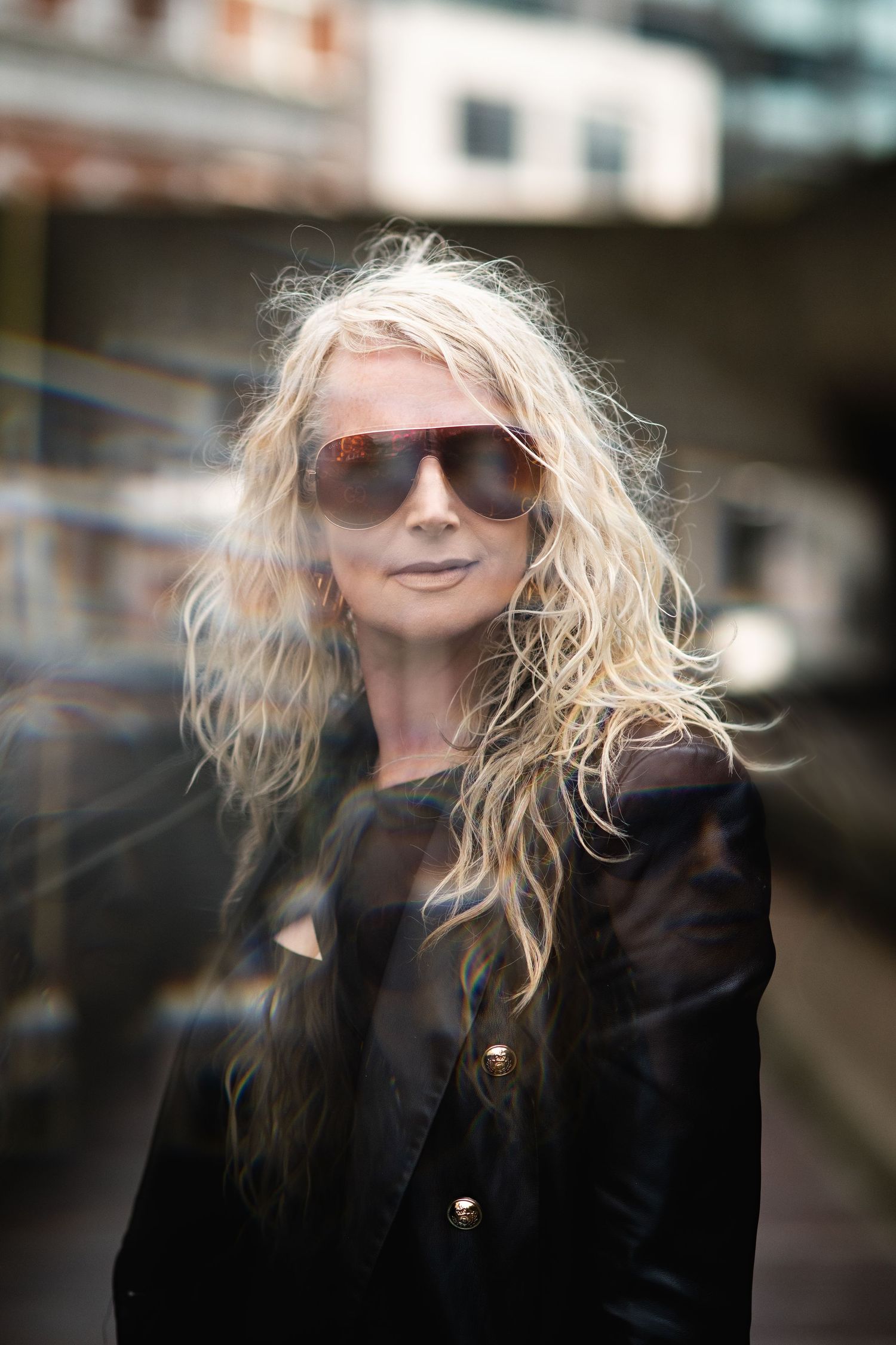 Blonde woman with wavy hair wearing sunglasses and black jacket in an urban outdoor setting.