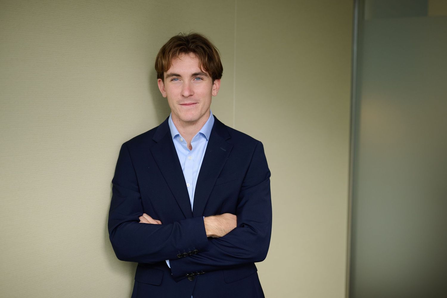 Smiling man in navy suit with arms crossed leaning against neutral-colored wall in professional setting.