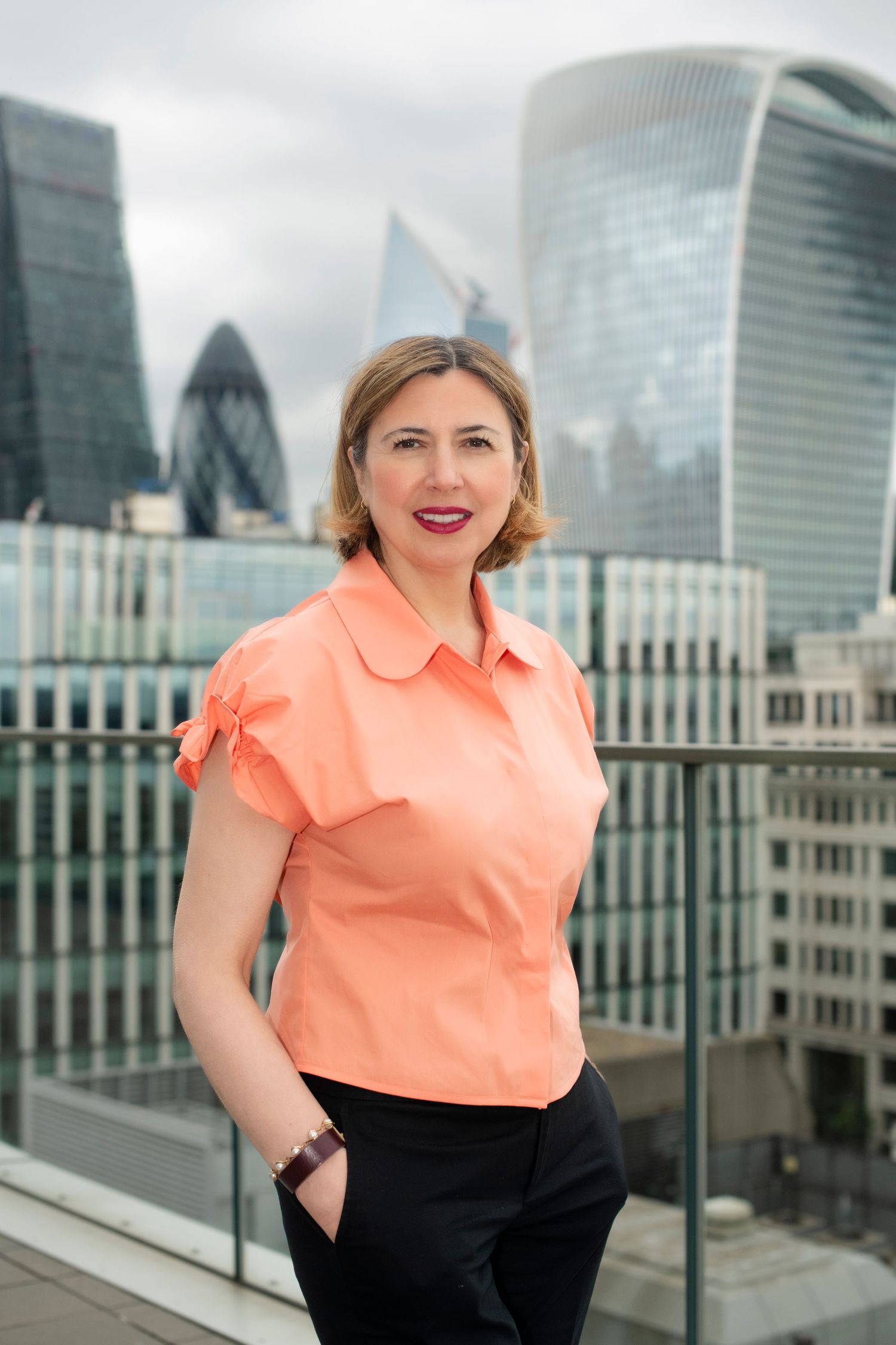 Woman in coral blouse standing on rooftop with London skyline behind her.