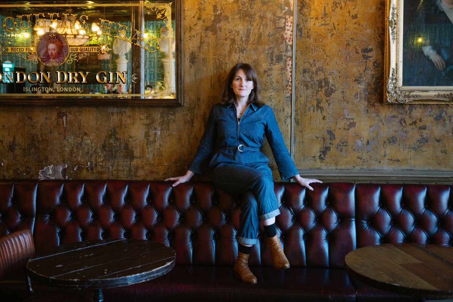 Woman in denim jumpsuit sitting on leather sofa in vintage bar with London Dry Gin sign.
