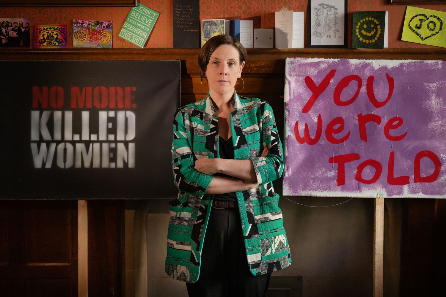 Woman in green patterned blazer stands before protest signs reading No More Killed Women and You Were Told.