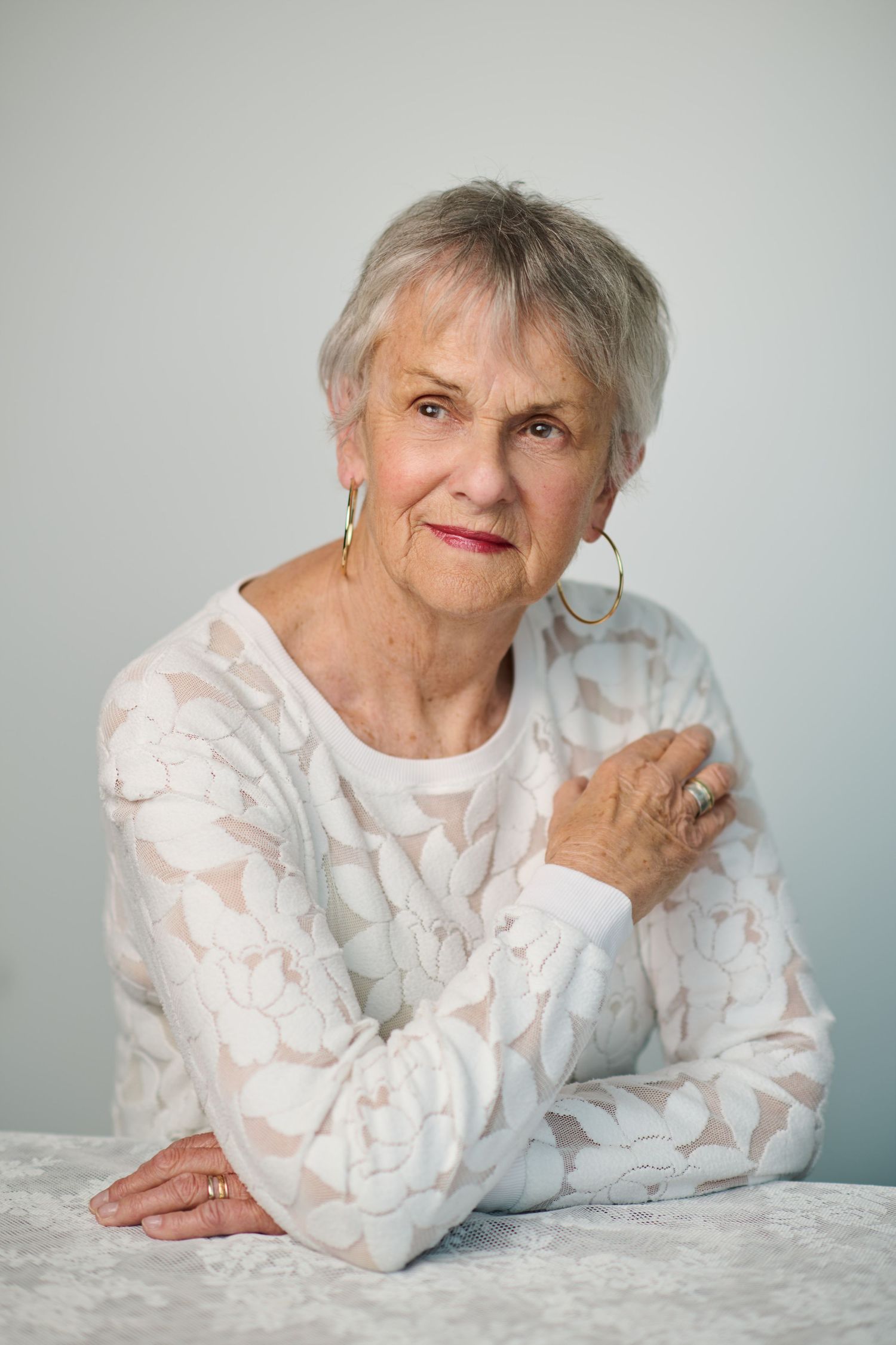 Elegant senior woman with short gray hair wearing a white lace top and gold hoop earrings, posing gracefully at a table.