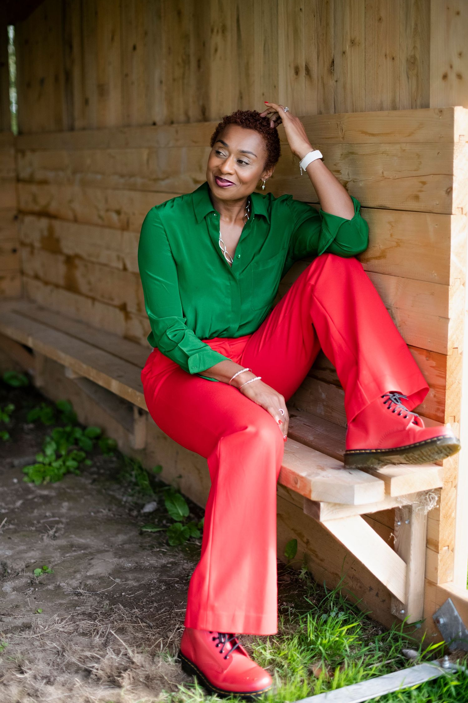 Black woman in green shirt and red pants sitting relaxed on a wooden bench in an outdoor shelter.