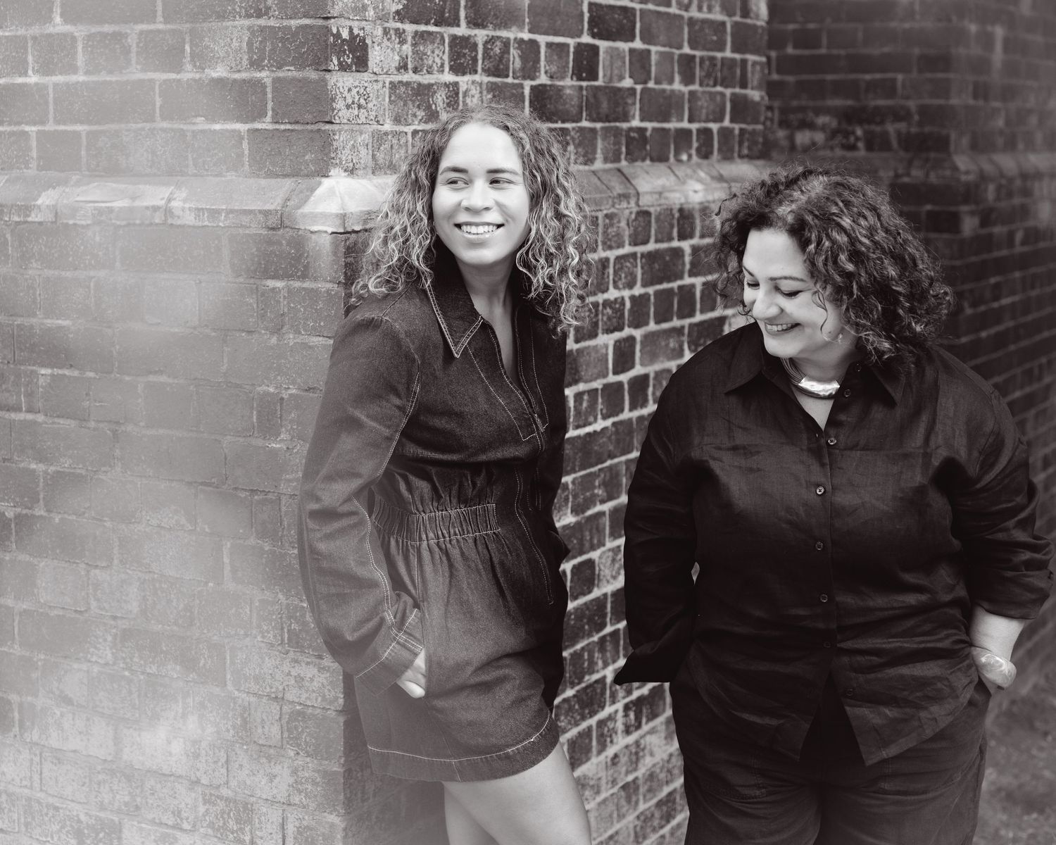 Two curly-haired women smile and laugh together leaning against a brick wall in a black and white photo.