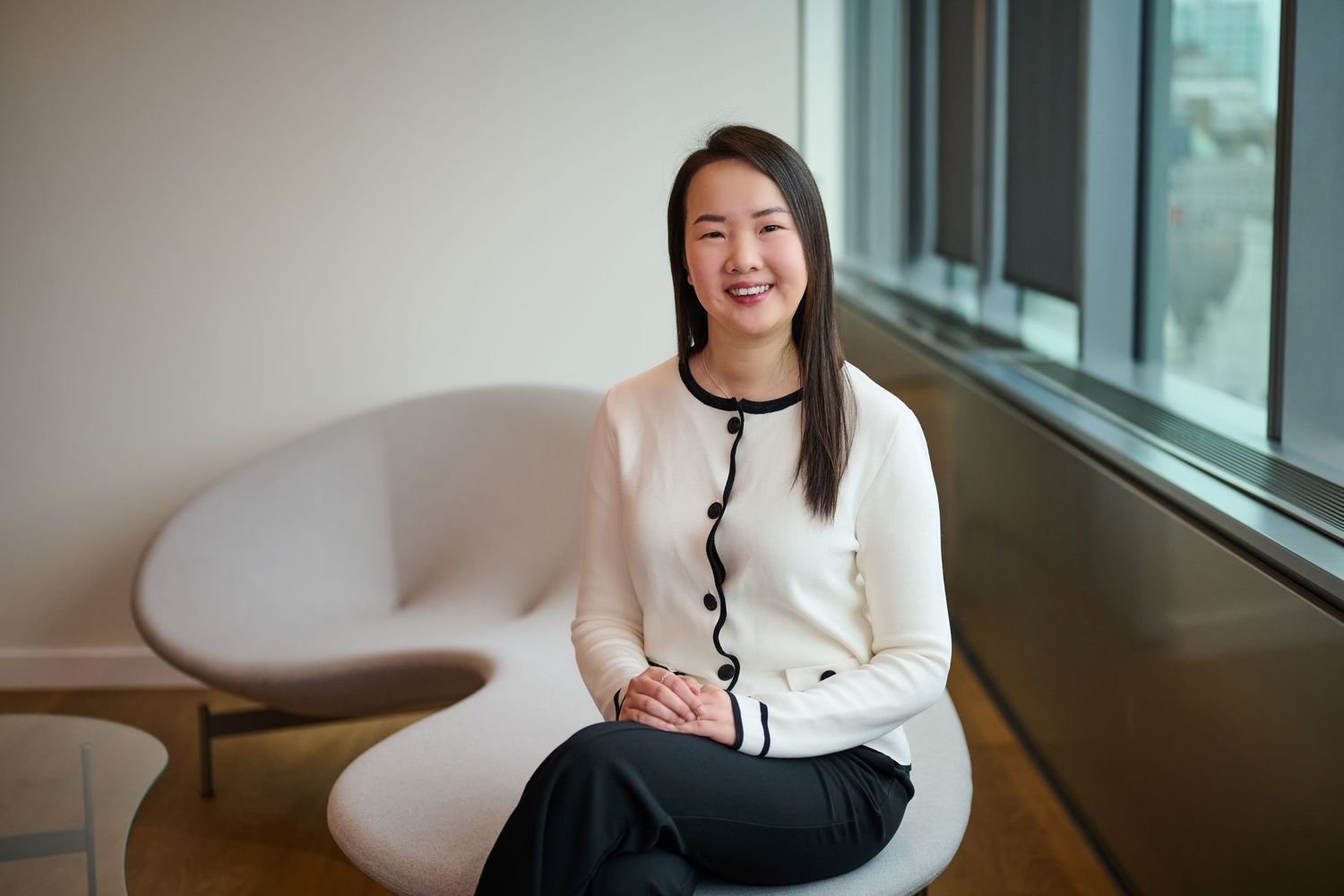 Young Asian woman smiling in modern office, wearing white cardigan and black pants, seated in white chair by window.