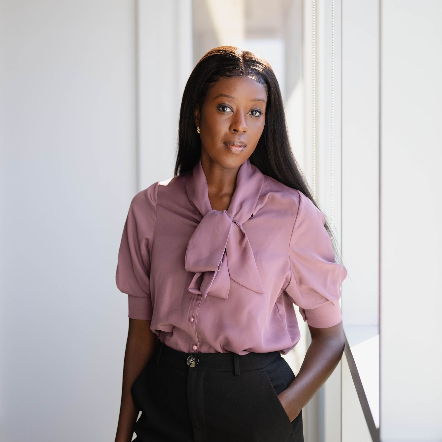 Young woman in mauve bow blouse and black pants posing near bright window indoors.