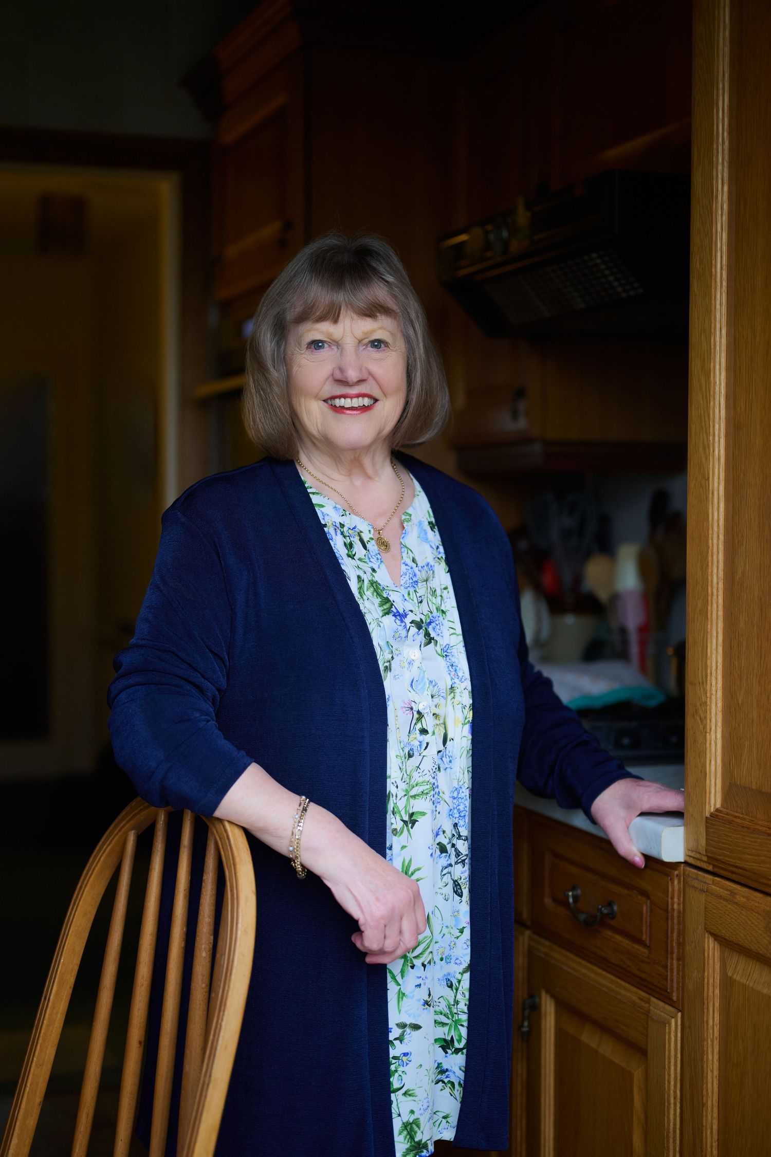 Smiling older woman in blue cardigan and floral dress stands in cozy kitchen doorway.