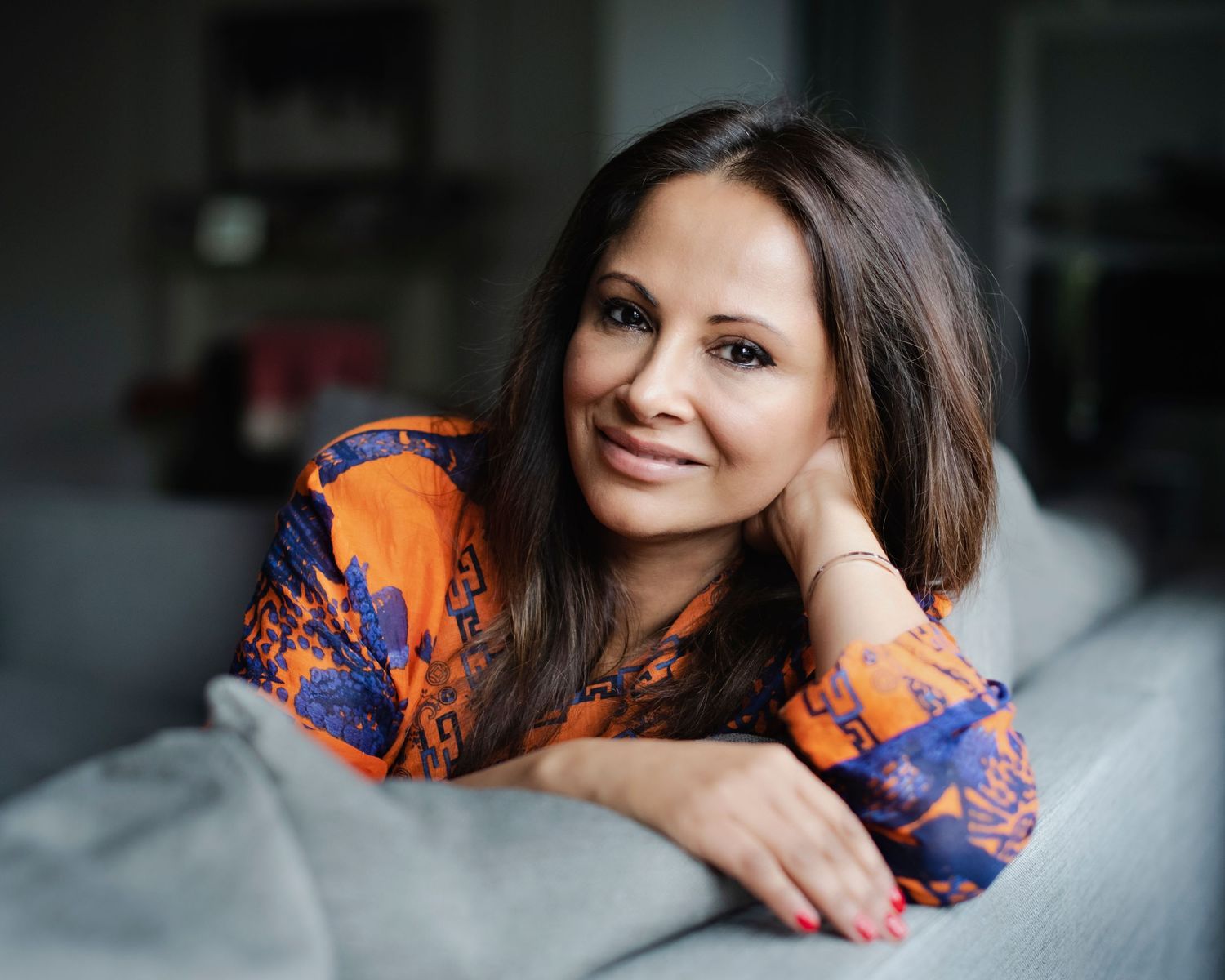 Smiling woman in colorful orange top relaxes on grey sofa in modern living room.