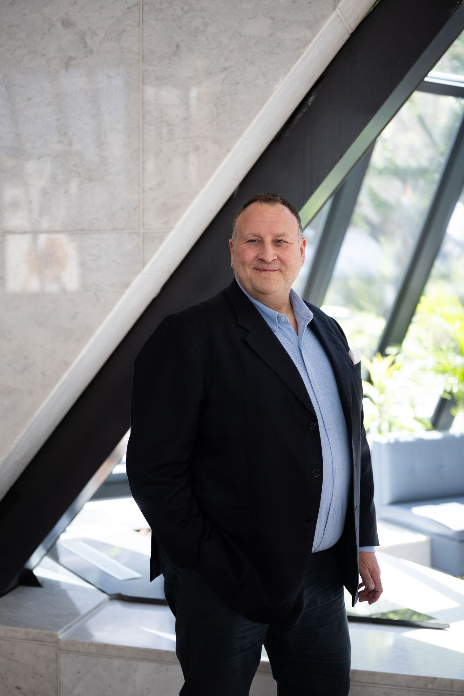 Smiling man in black blazer and light blue shirt posing near angular concrete architecture with natural light.