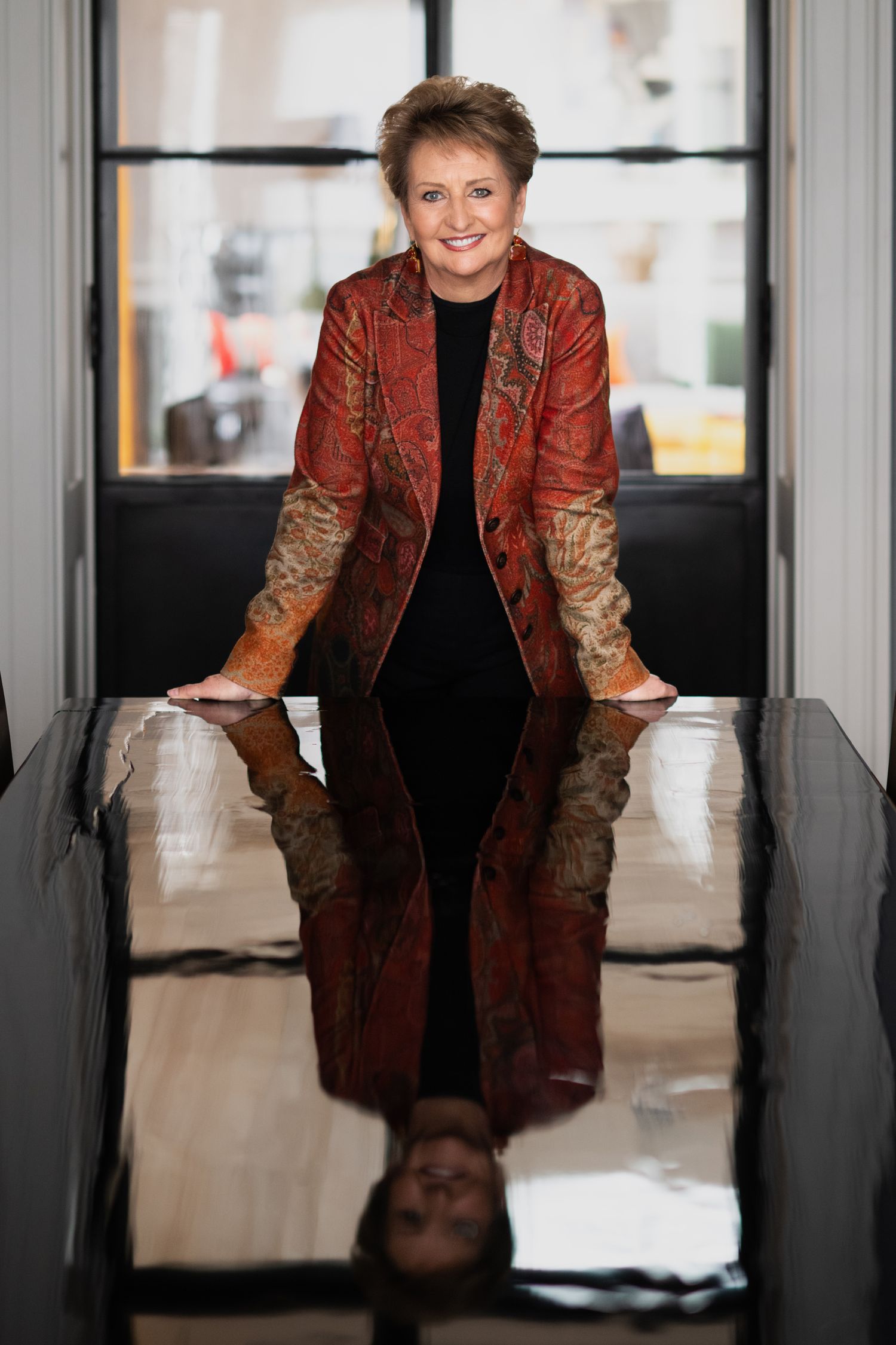 Smiling woman in red paisley jacket leaning on reflective black table in modern space.