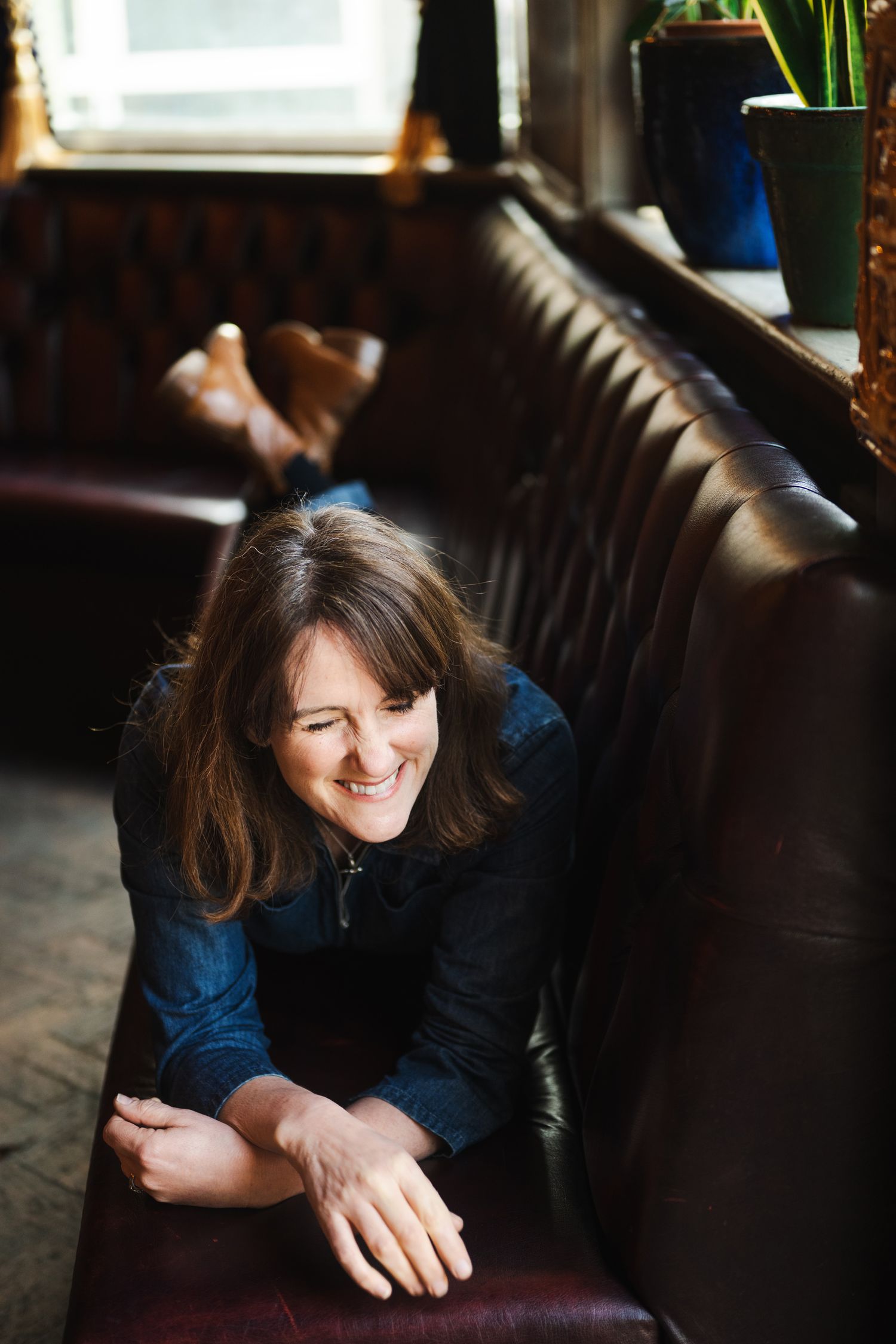 Happy brunette woman leaning on leather pub sofa, smiling downward in warm light.