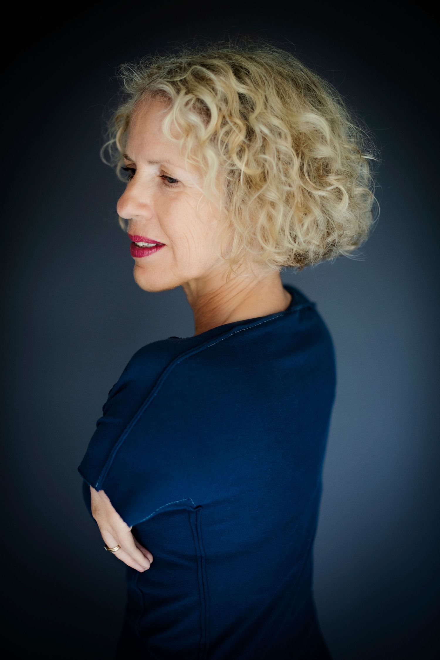A side profile of a curly blonde-haired woman in a navy top with arms crossed against a dark background.