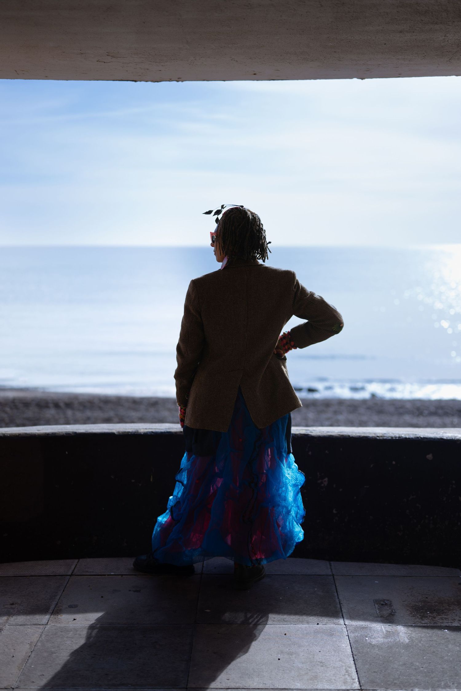 A woman in a blue skirt and brown jacket stands silhouetted in an archway overlooking the sea.