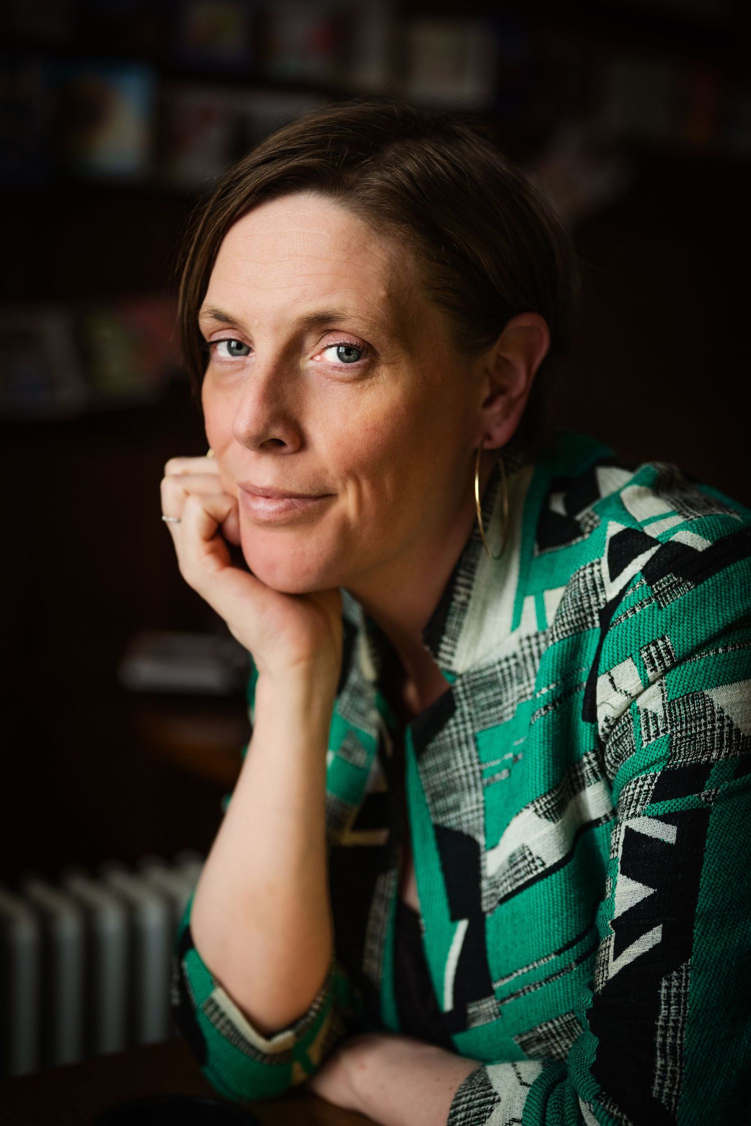 Woman in green geometric blazer rests chin on hand, gazing thoughtfully in a warmly lit room.