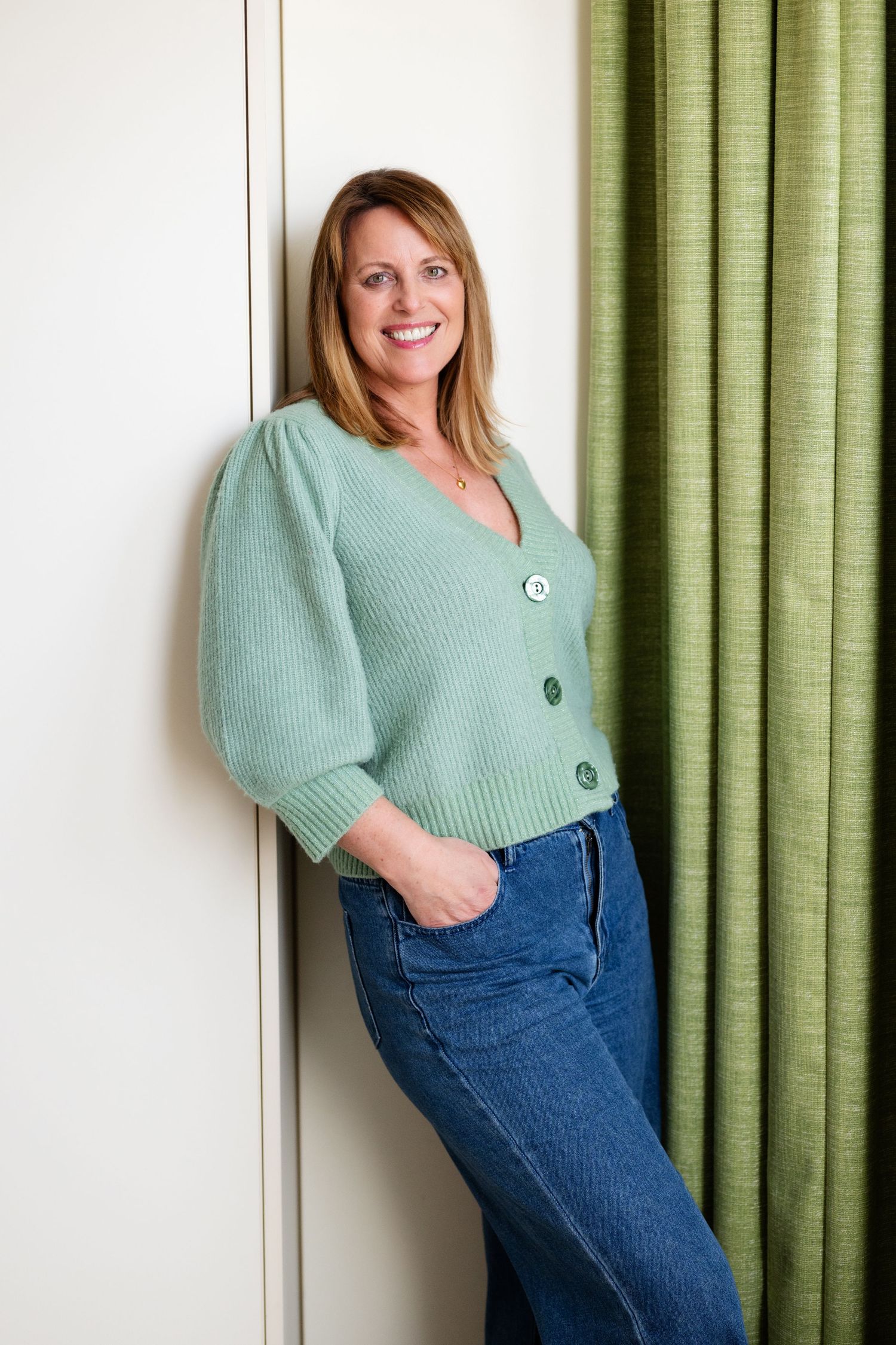 Smiling blonde woman in mint green knit cardigan and jeans leaning against white wall near green curtain.
