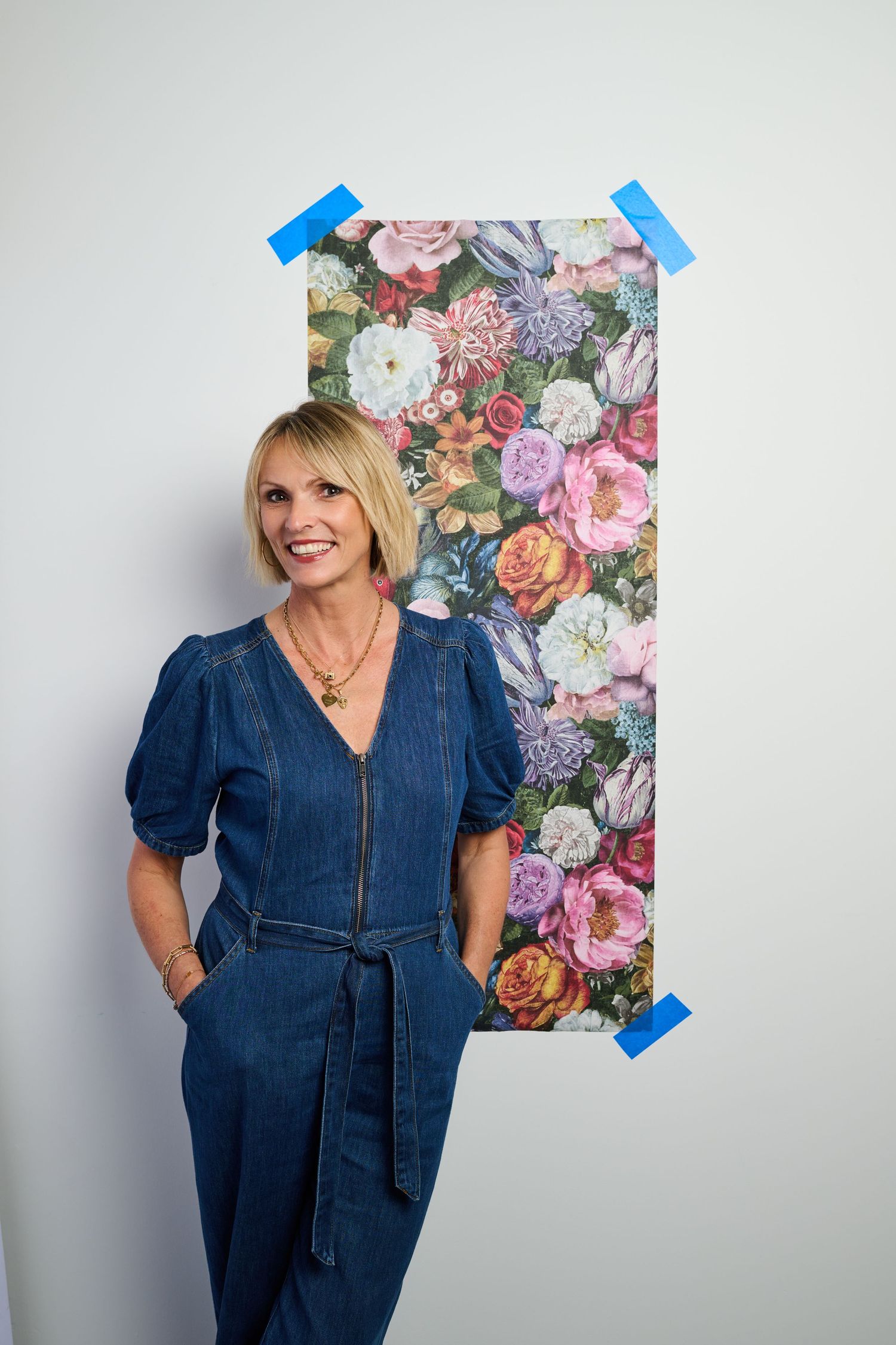 Smiling blonde woman in denim jumpsuit standing against white wall with colorful floral wallpaper sample taped behind her.