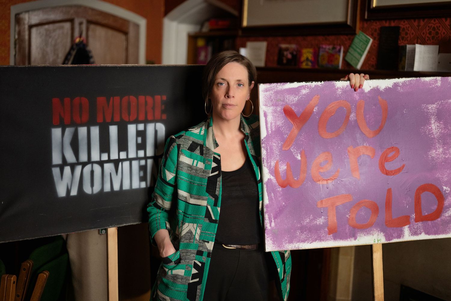 Woman standing between protest signs reading No More Killed Women and You Were Told.
