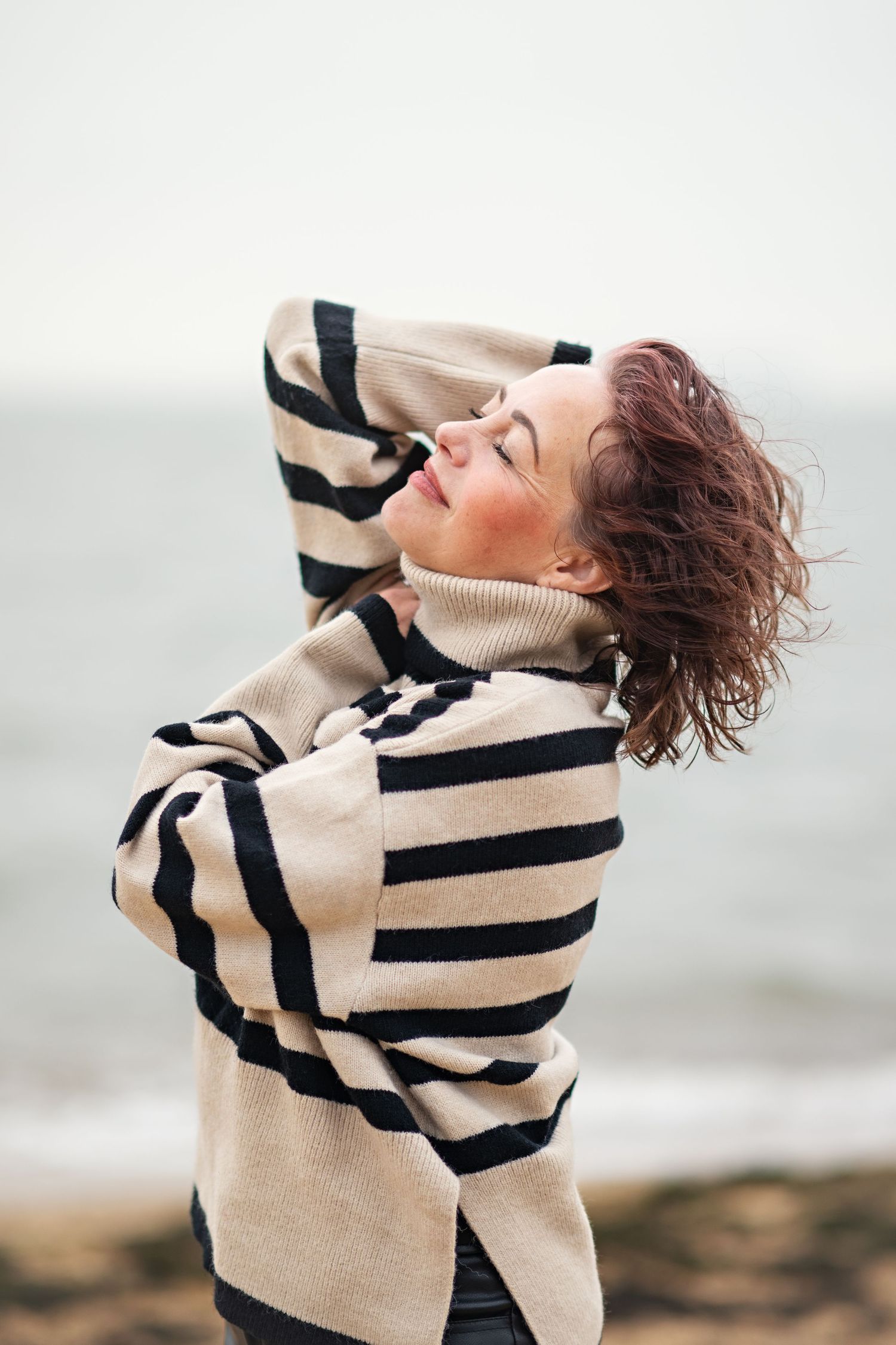Woman in striped turtleneck sweater tilts head back with eyes closed on a cloudy beach.