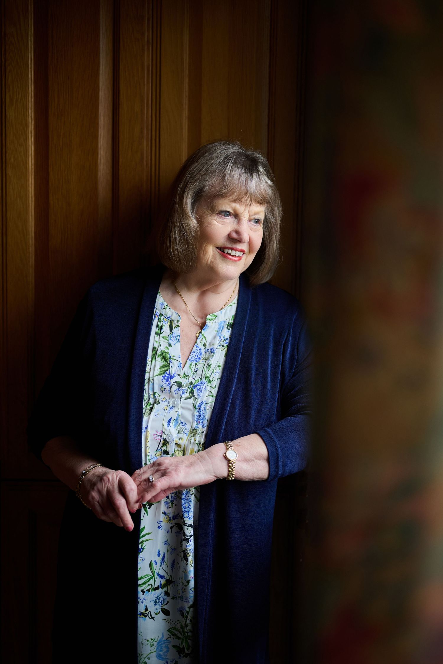 Cheerful senior woman in blue cardigan and floral dress leans against wooden wall smiling warmly.