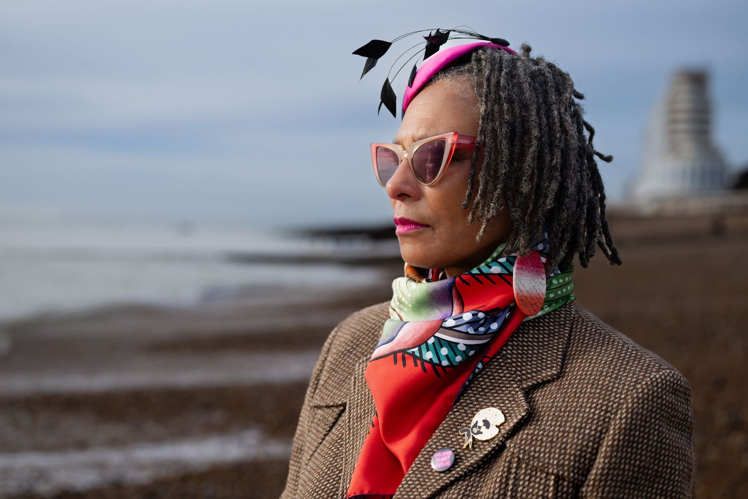 An elegant woman with gray locs wearing cat-eye sunglasses and colorful scarf stands on a pebble beach.