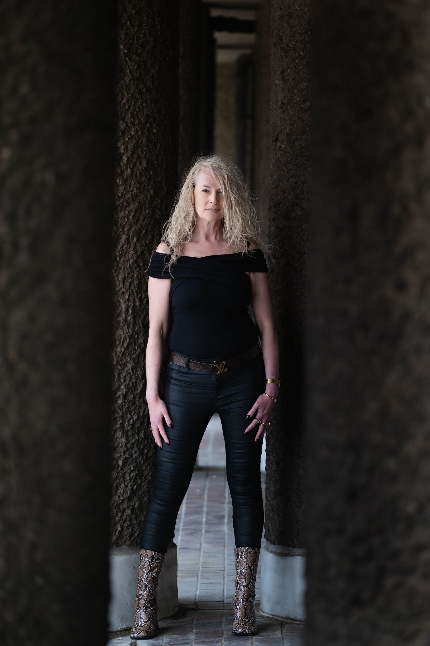 Blonde woman in black off-shoulder top, leather pants, and snake-print boots standing between stone columns.