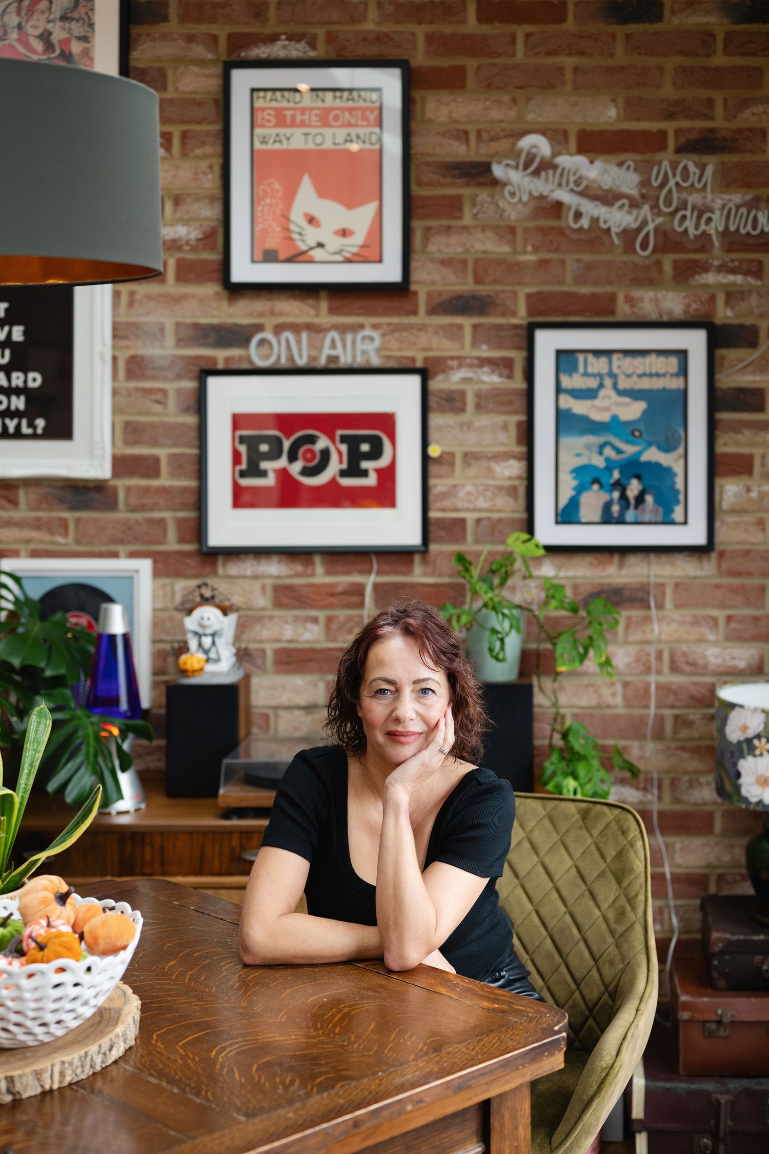 Brunette woman sitting at wooden table in front of eclectic brick wall with framed posters.