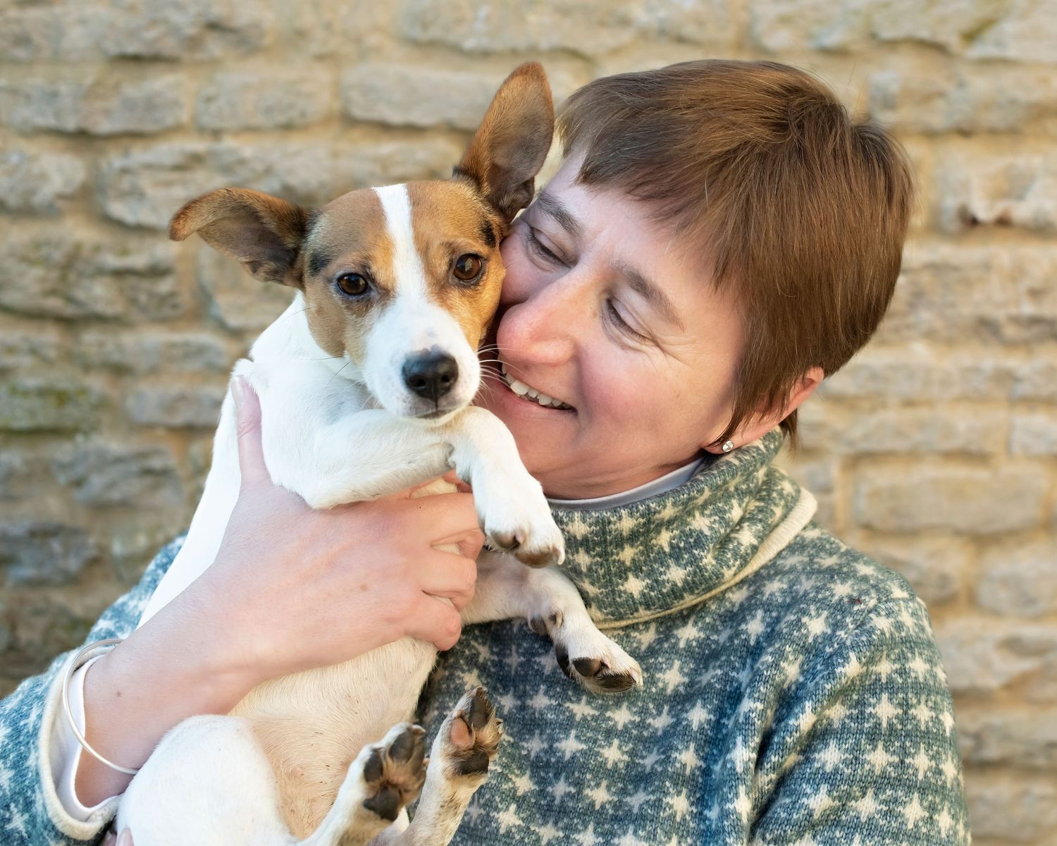 Smiling woman in a green patterned sweater holding a small Jack Russell terrier dog against a stone wall.