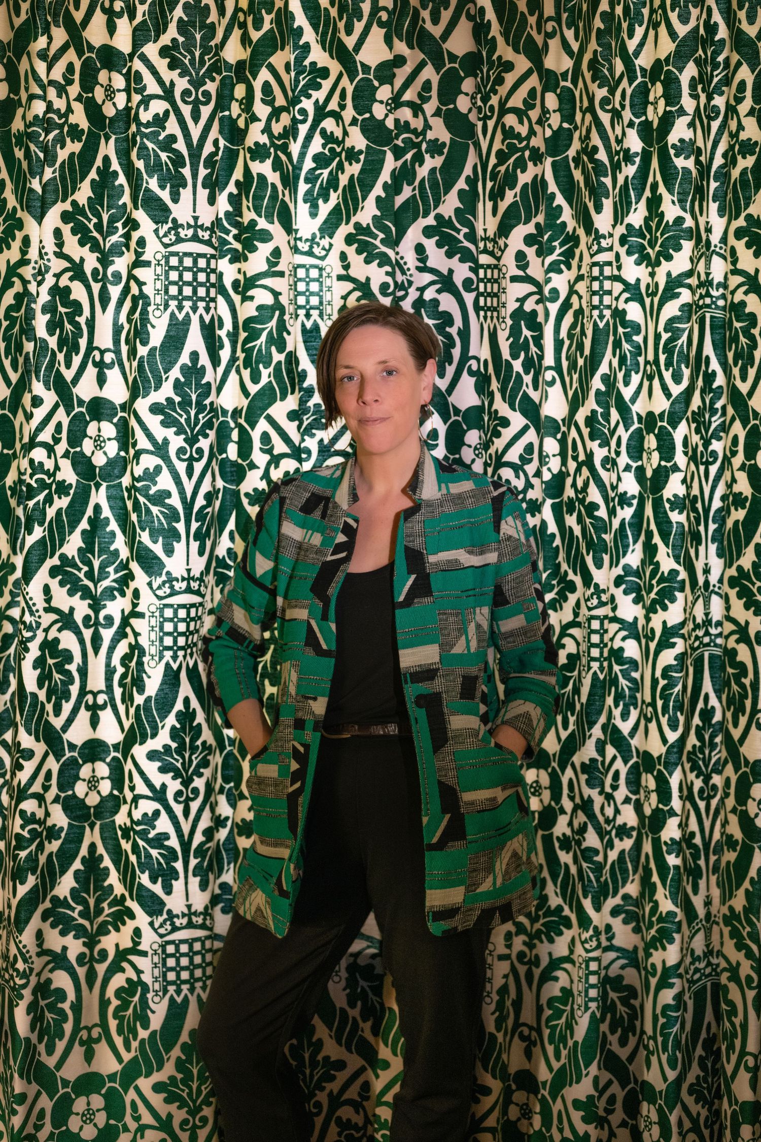 Woman in green patterned blazer poses confidently against ornate green and white damask wallpaper.