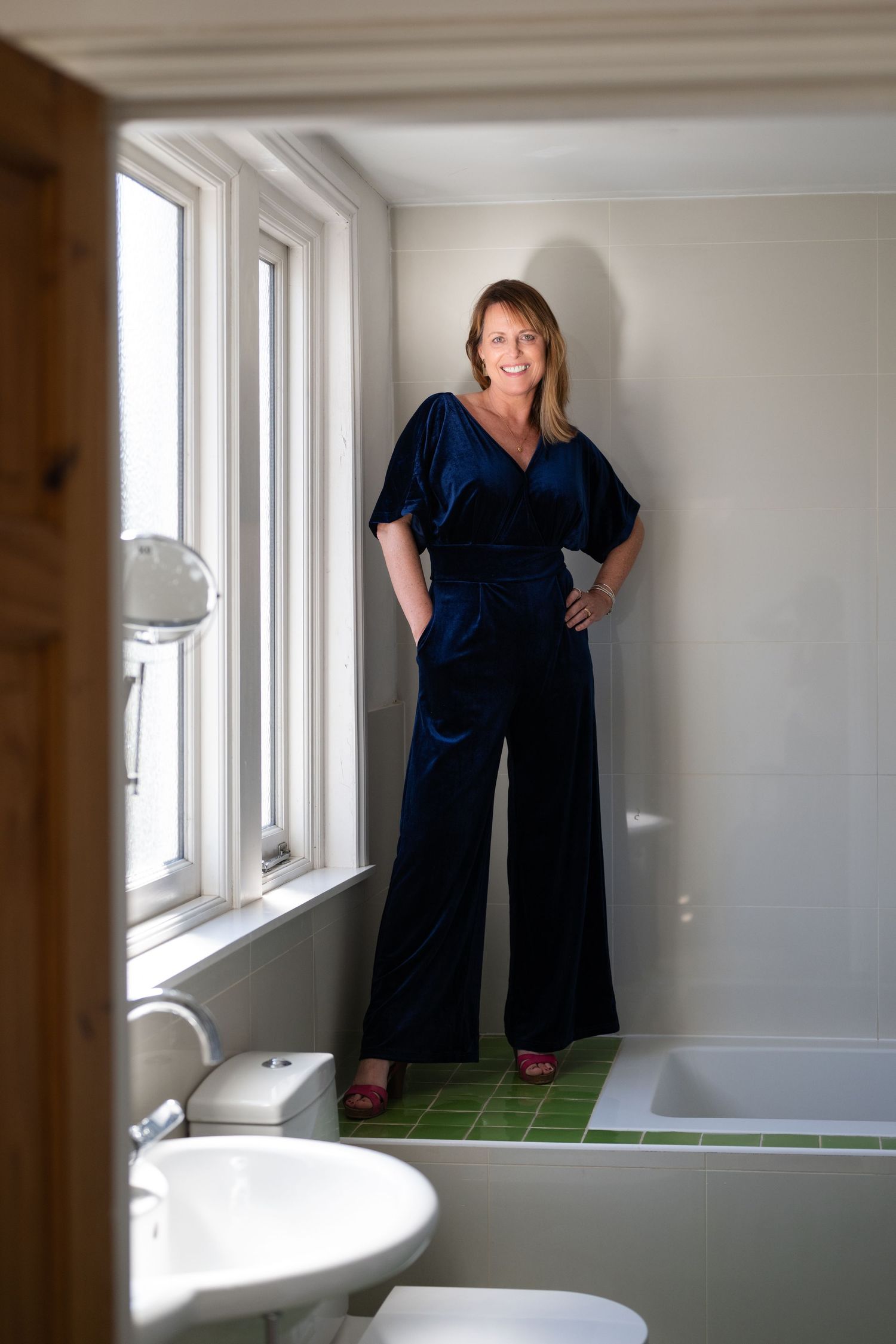 Smiling woman wearing elegant navy velvet wide-leg jumpsuit posing in modern white bathroom by window.