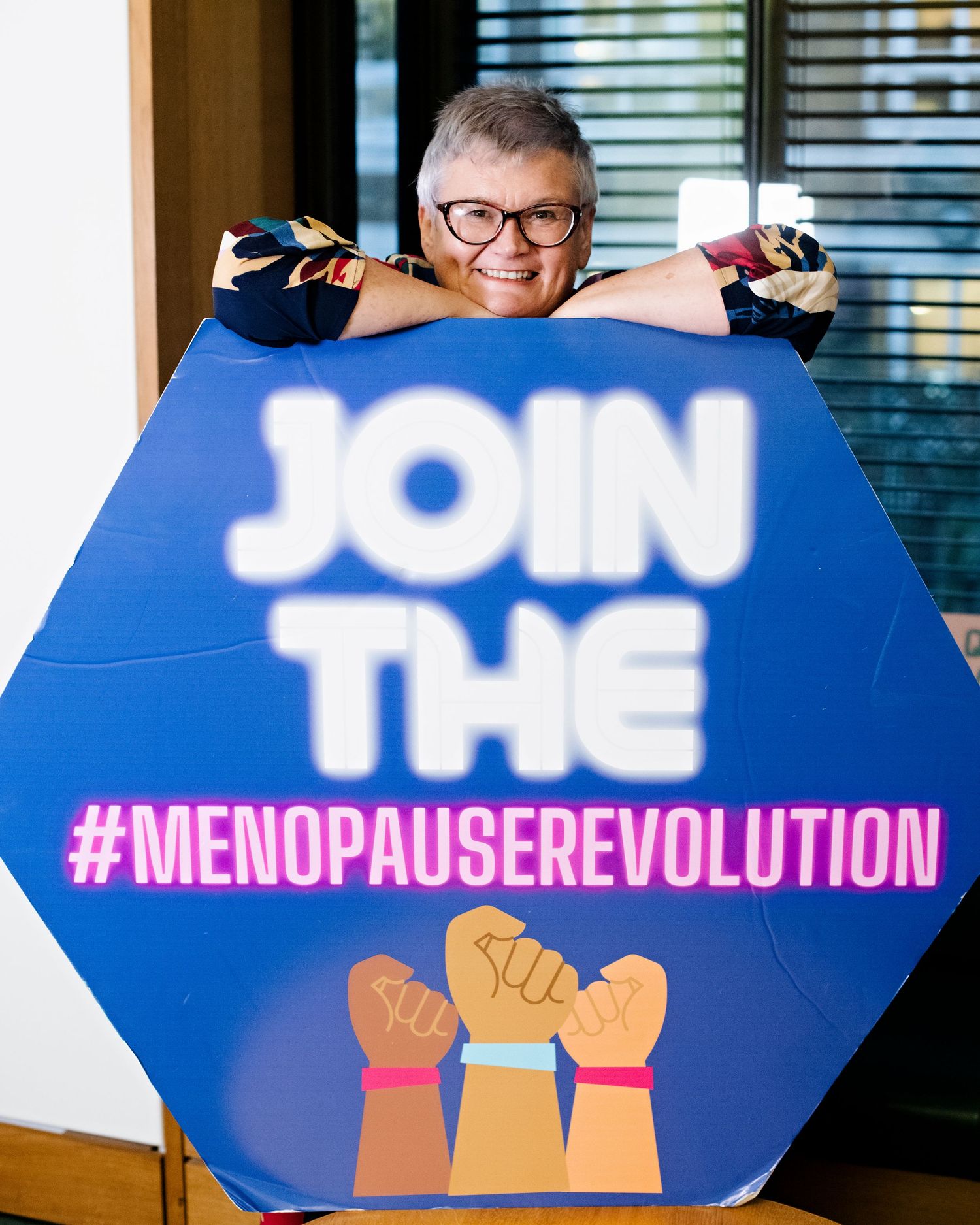 Smiling woman with gray hair and glasses posing behind a blue menopause revolution awareness campaign sign.
