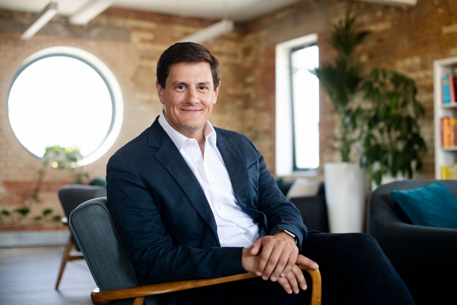 Smiling businessman in navy suit seated in modern office with brick walls.