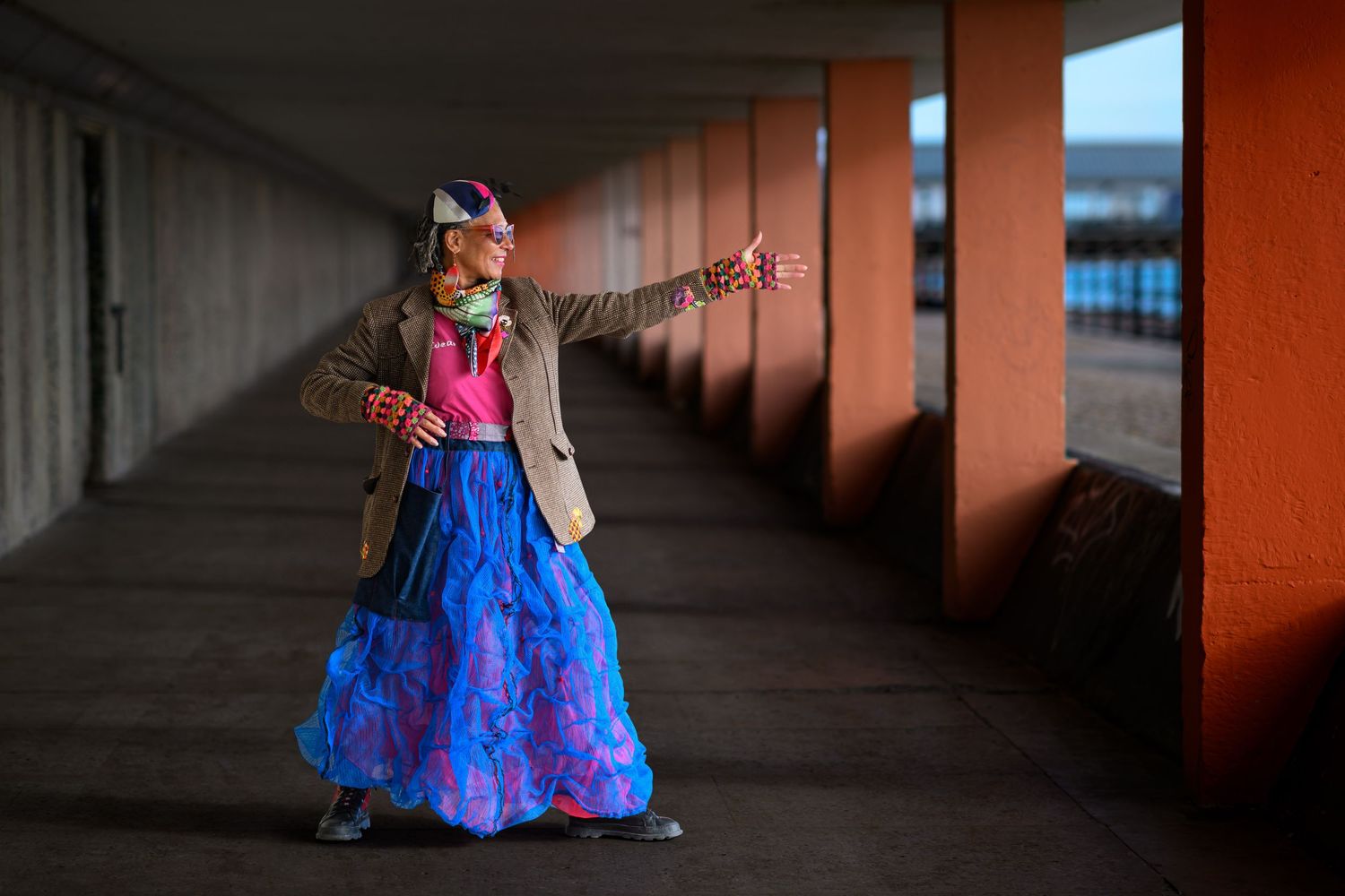 Person in colorful blue skirt and floral mask posing in urban corridor with orange pillars.