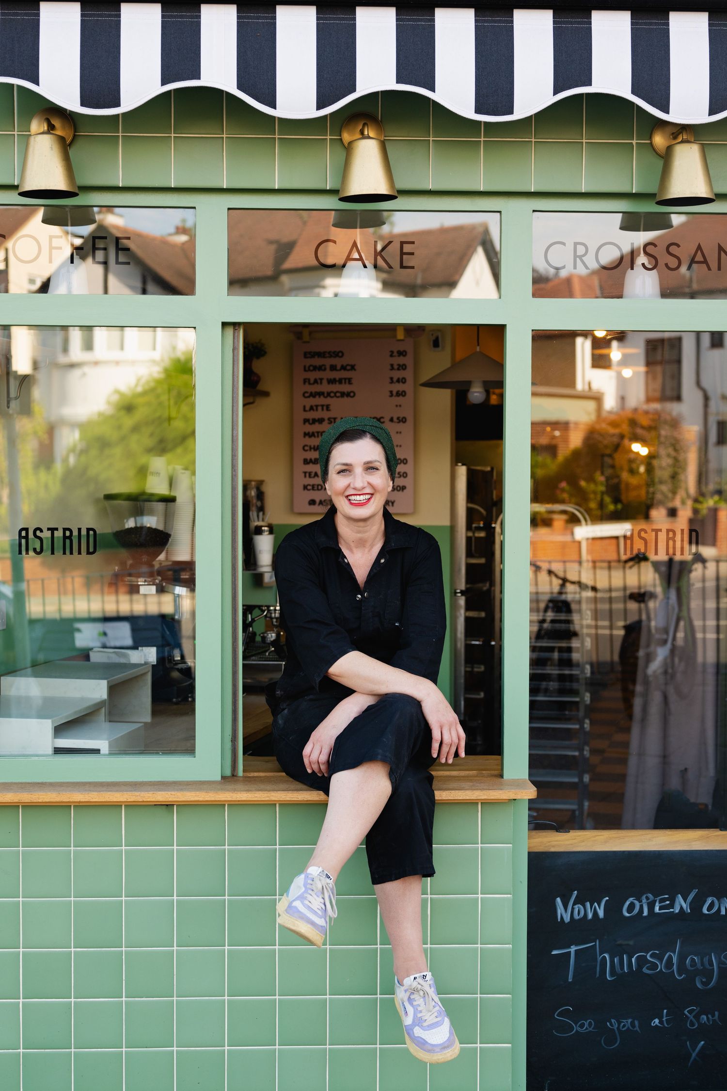 Smiling woman with green hair sits on the window ledge of a charming green cafe storefront.