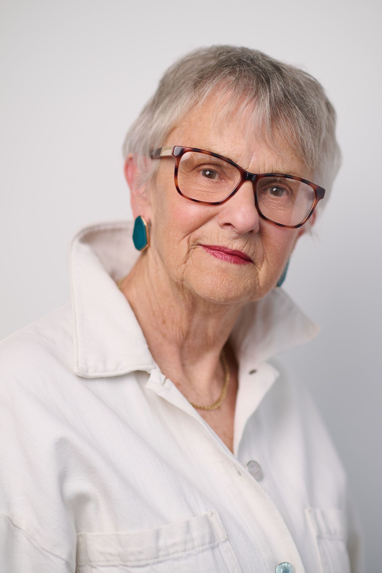 Elderly woman with short gray hair, red glasses, and teal earrings wearing a white jacket against a light background.