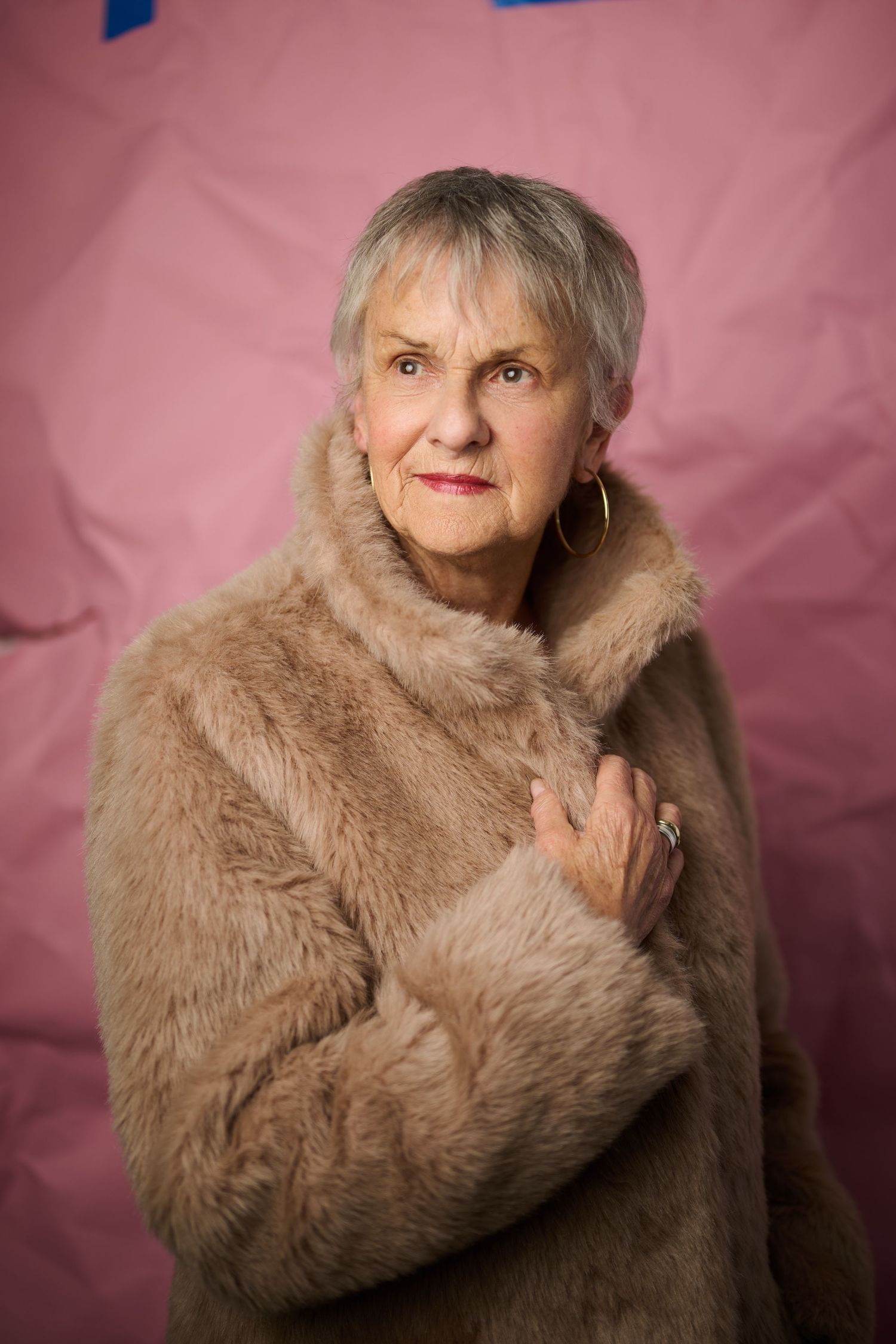 Elegant senior woman in beige fur coat posing against pink background, wearing gold hoop earrings.
