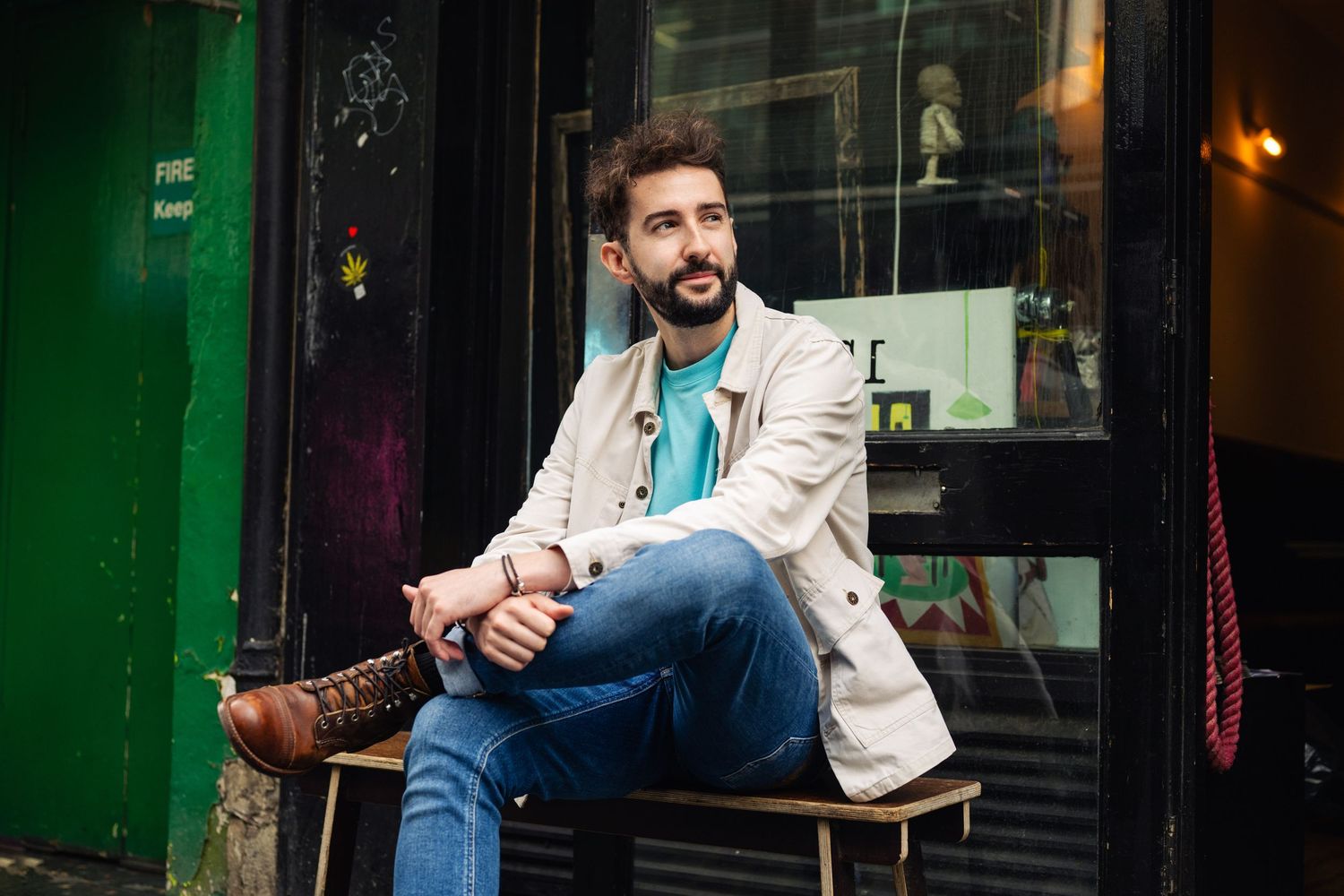 Bearded man in casual jacket and jeans sitting outside a colorful urban storefront, looking thoughtfully to the side.