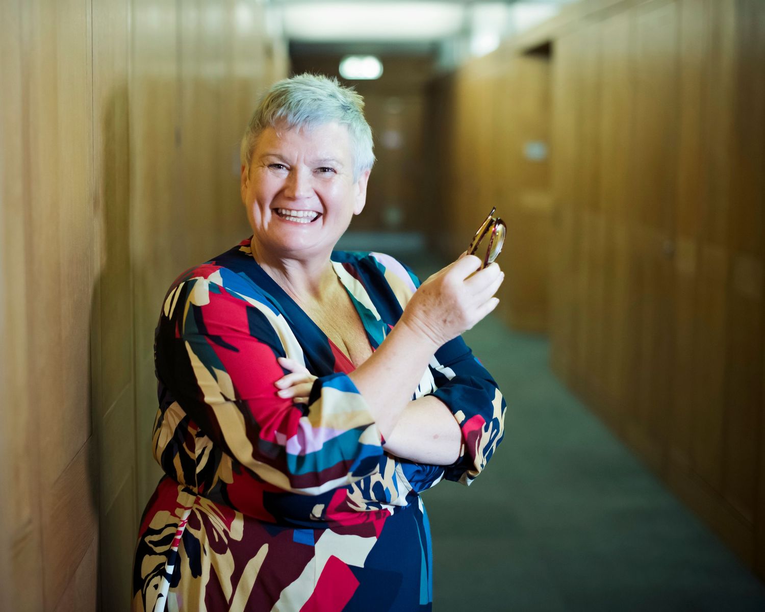 Cheerful older woman with short gray hair smiling while holding glasses and leaning against a wooden wall.