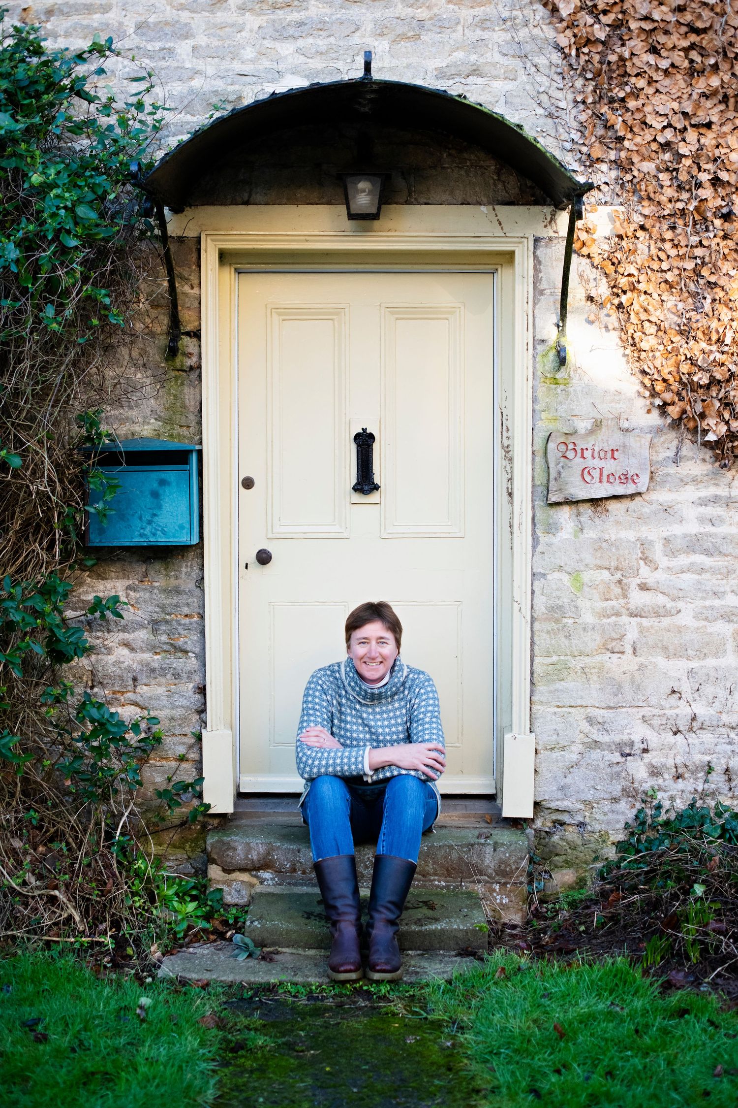 Smiling woman sitting on stone steps in front of a cream-colored door of a rustic cottage.
