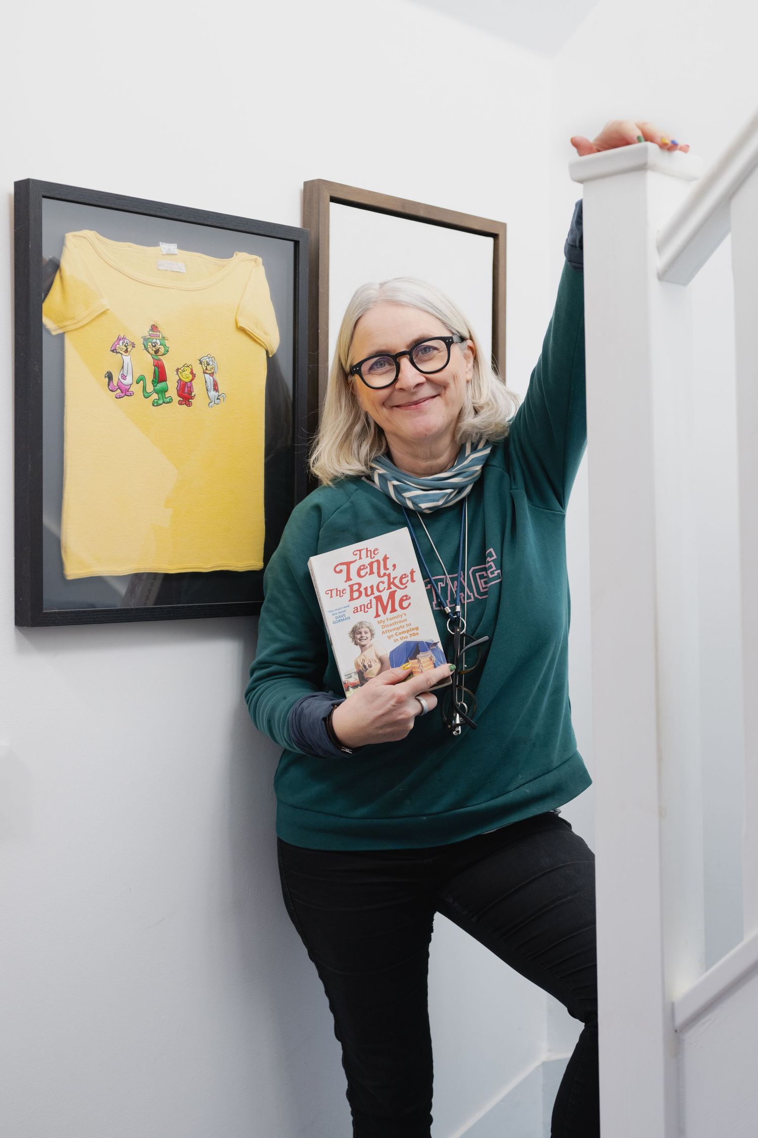 Woman with gray bob holds a book beside framed yellow shirt artwork on a white wall.