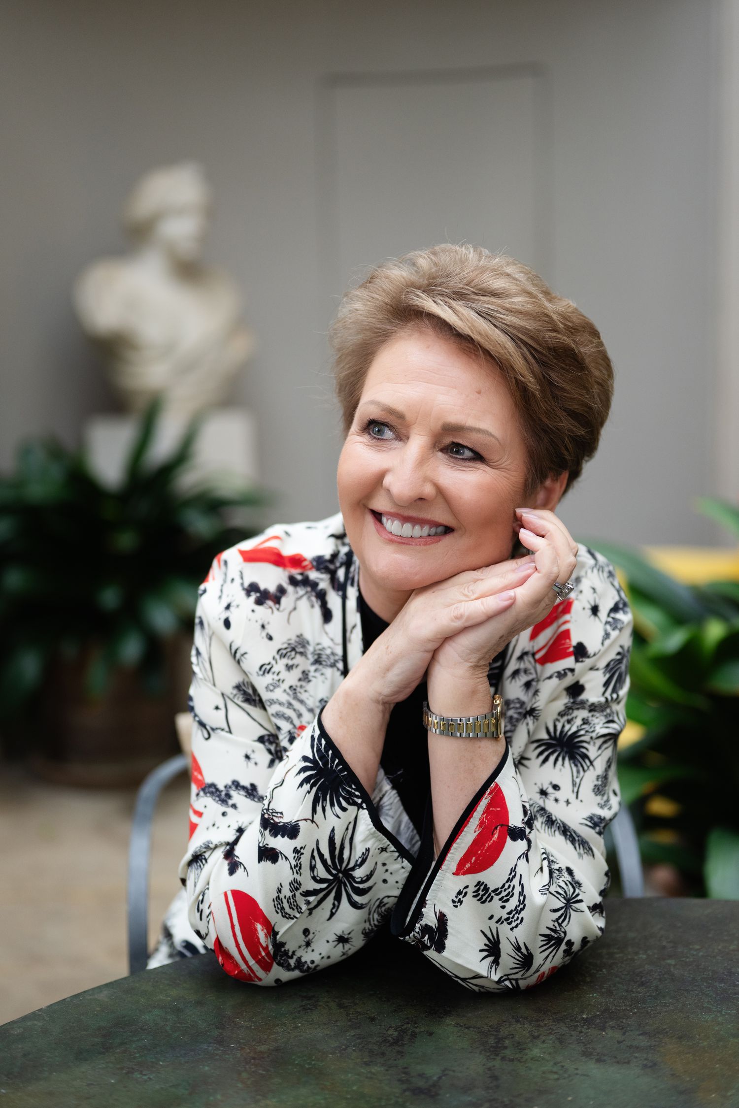Smiling woman in floral blazer resting chin on hands at table near classical sculpture.
