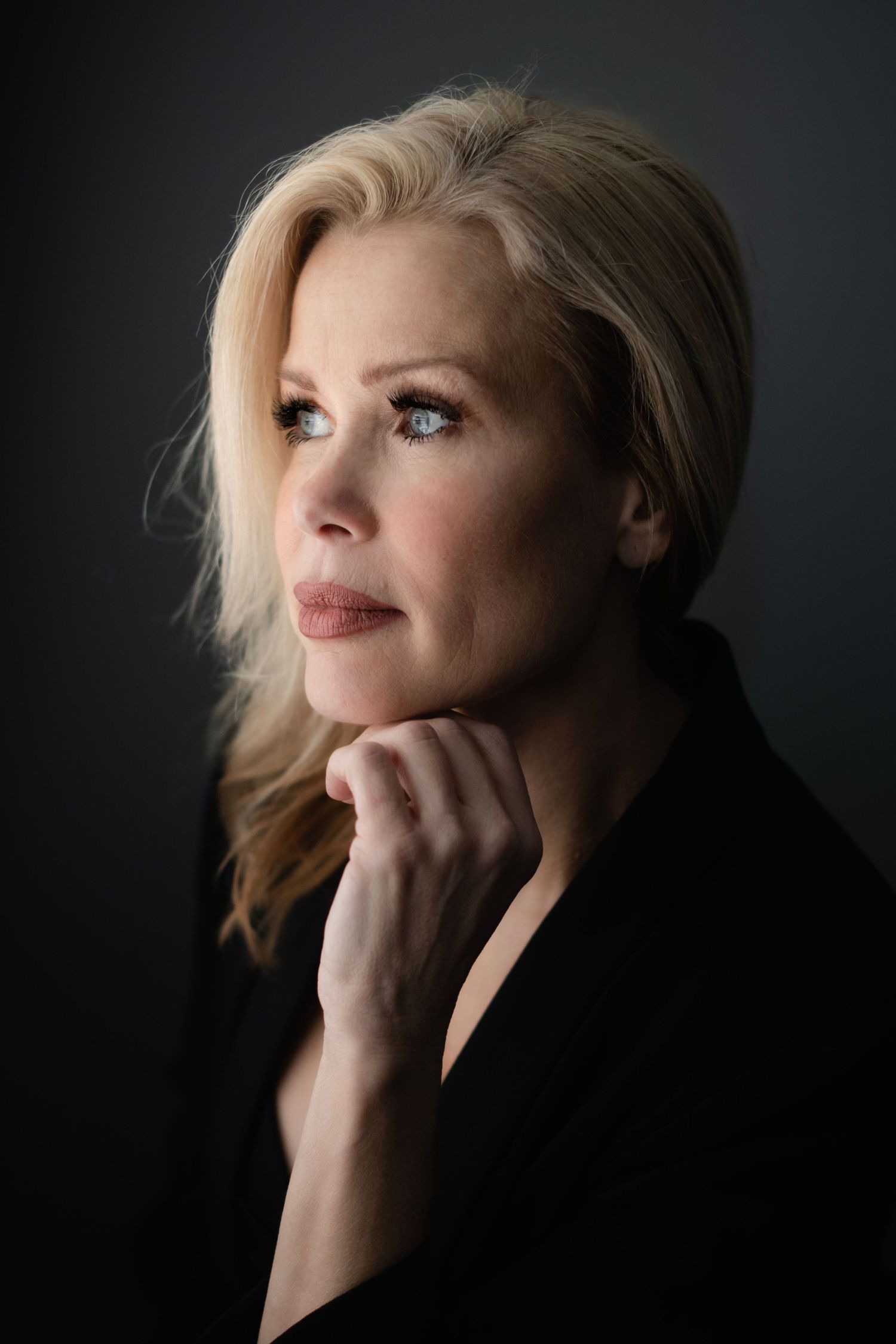 Elegant blonde woman in black jacket rests chin on hand, gazing thoughtfully in a professional studio portrait.