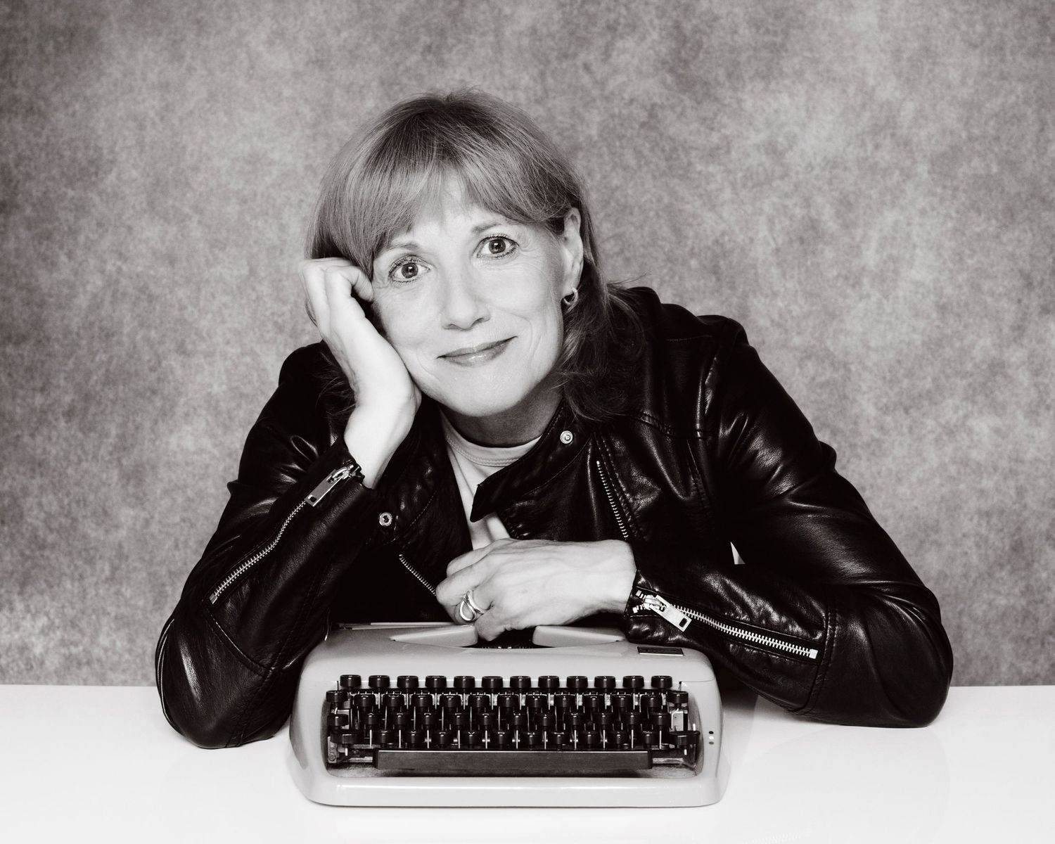 Woman in leather jacket poses thoughtfully beside a vintage typewriter in a black and white portrait photo.