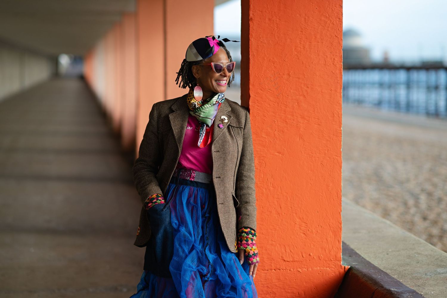 A stylish woman in a blue tulle skirt and tweed blazer poses by vibrant orange pillars near a beach.