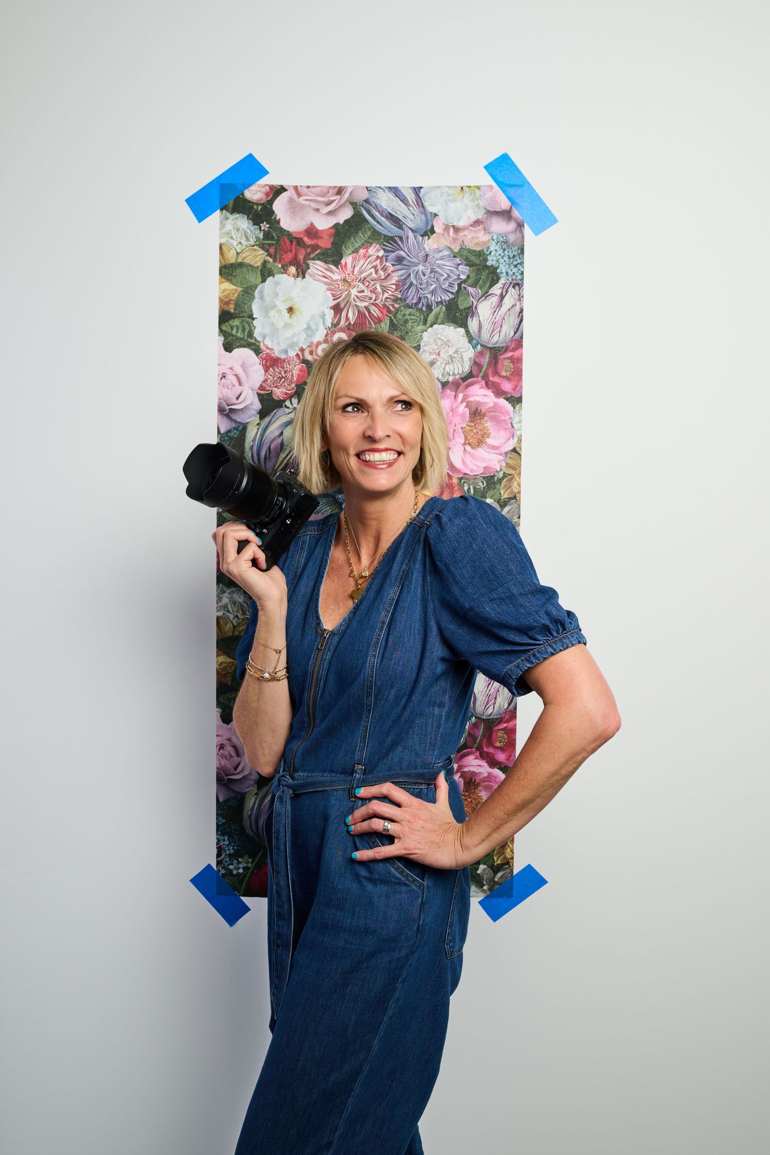 Smiling blonde woman in denim jumpsuit holding a camera with one hand on hip, posing before a floral poster on a white wall.
