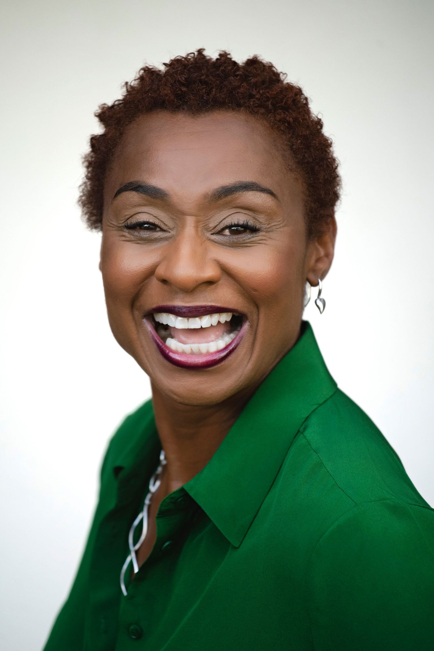 Smiling Black woman with short natural hair wearing a green shirt, laughing joyfully against a light background.