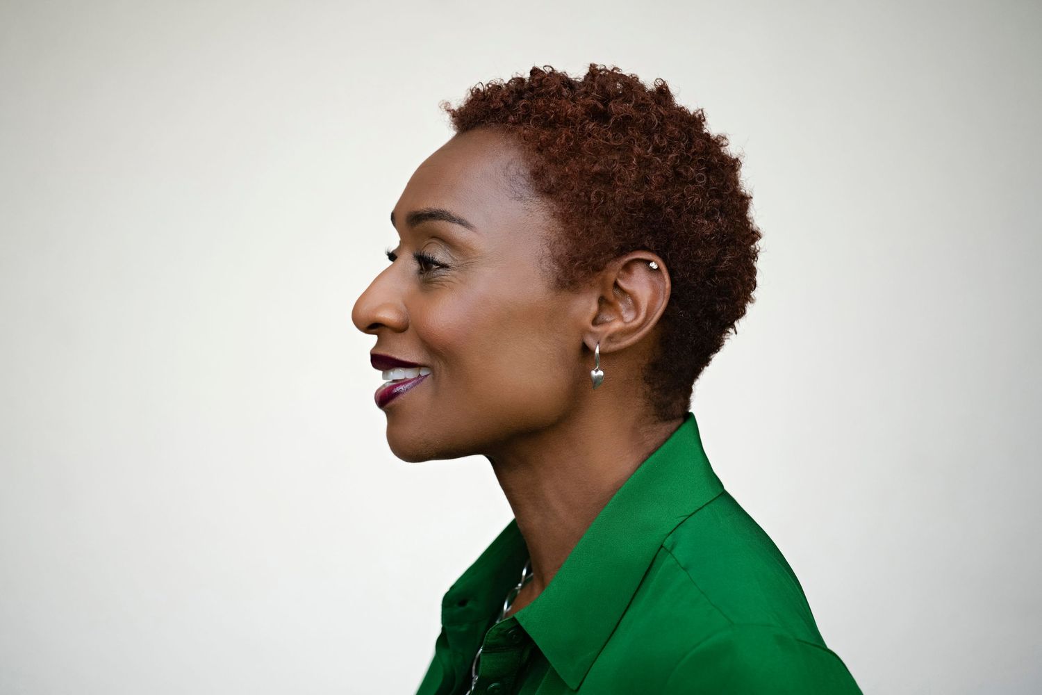 Side profile of a Black woman with short auburn hair wearing a green shirt and silver earring against white background.
