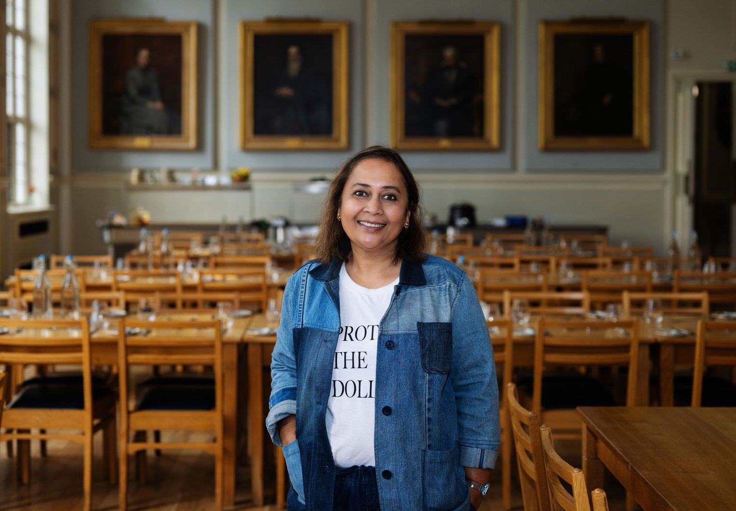 Woman in denim jacket and white graphic tee smiling in a formal dining hall with portraits.