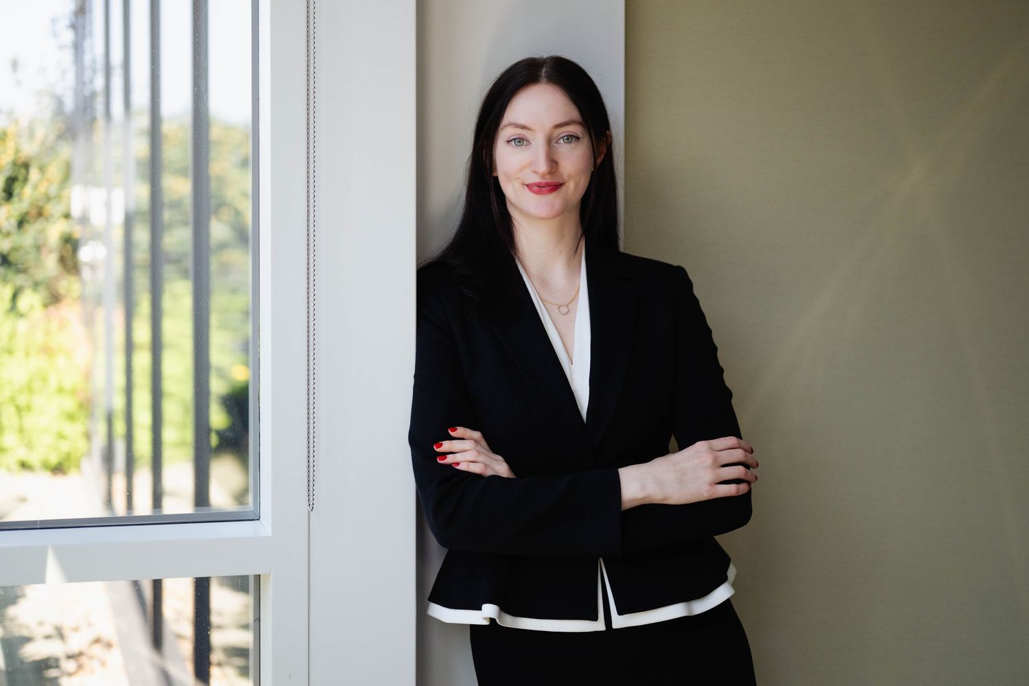 Professional woman in black blazer with arms crossed leaning against wall near window.