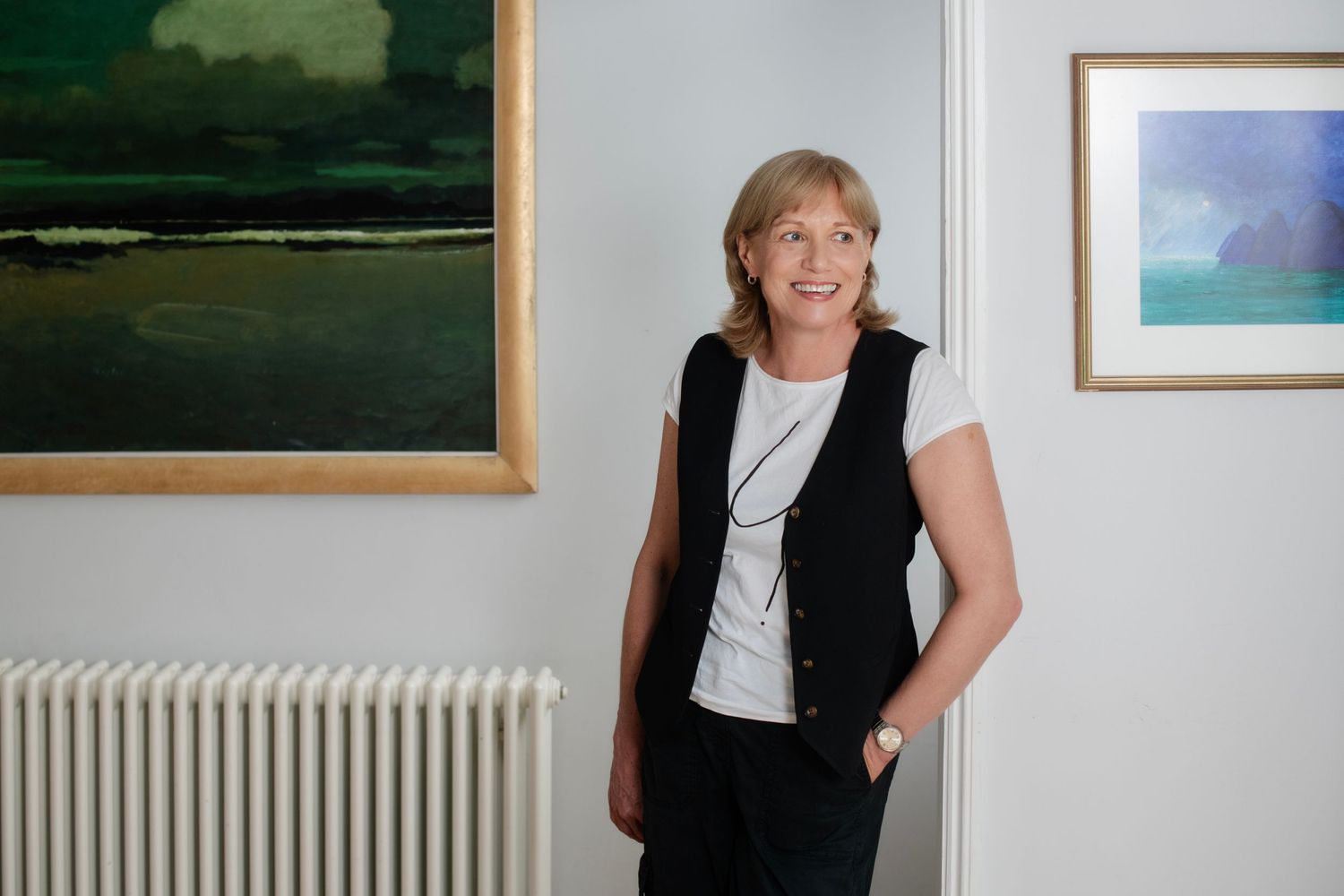 Smiling woman in black vest standing beside colorful landscape paintings in bright white gallery space.