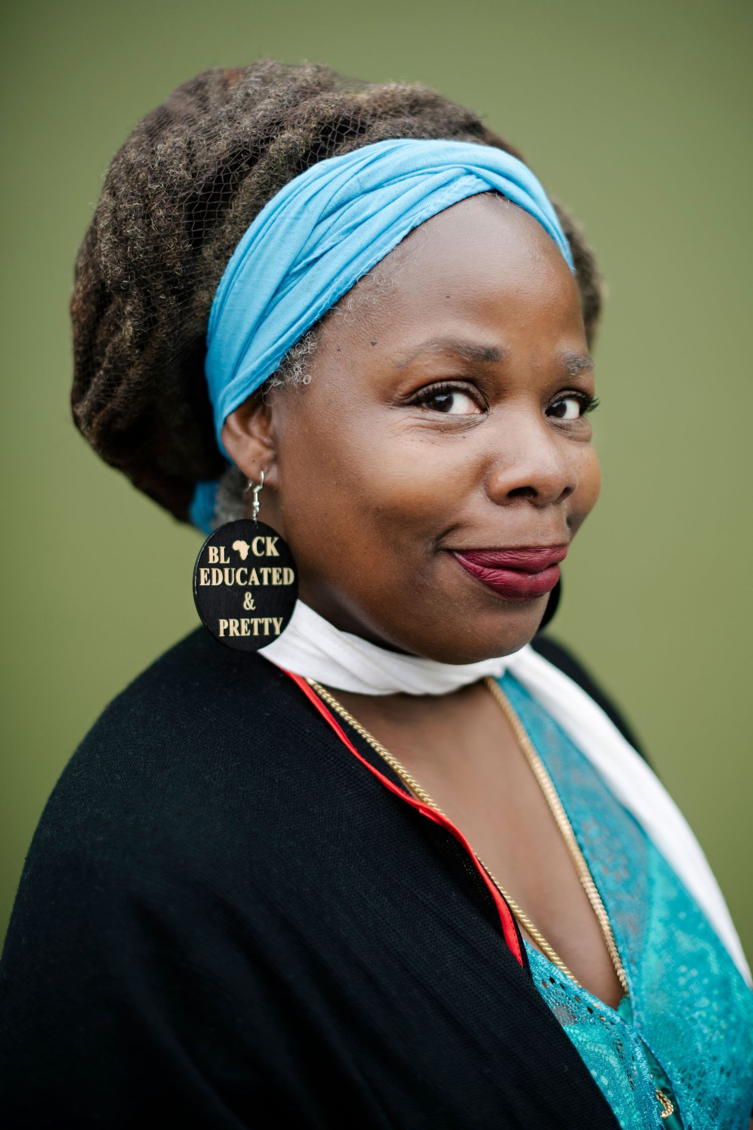 Close portrait of a woman with locs wearing Black Educated and Pretty earrings and a blue headband against green background.